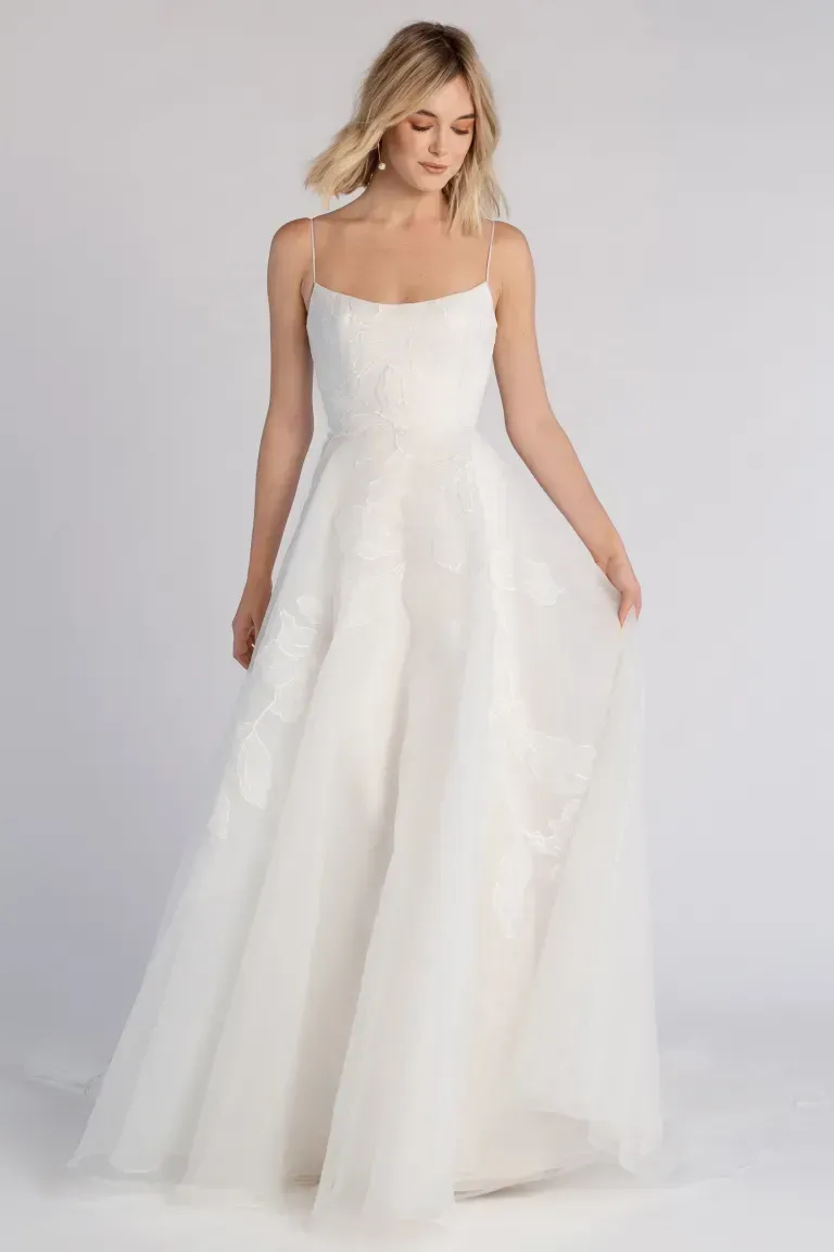 A person wearing a white, floor-length A-line wedding dress with spaghetti straps and a sheer, flowing tulle skirt.