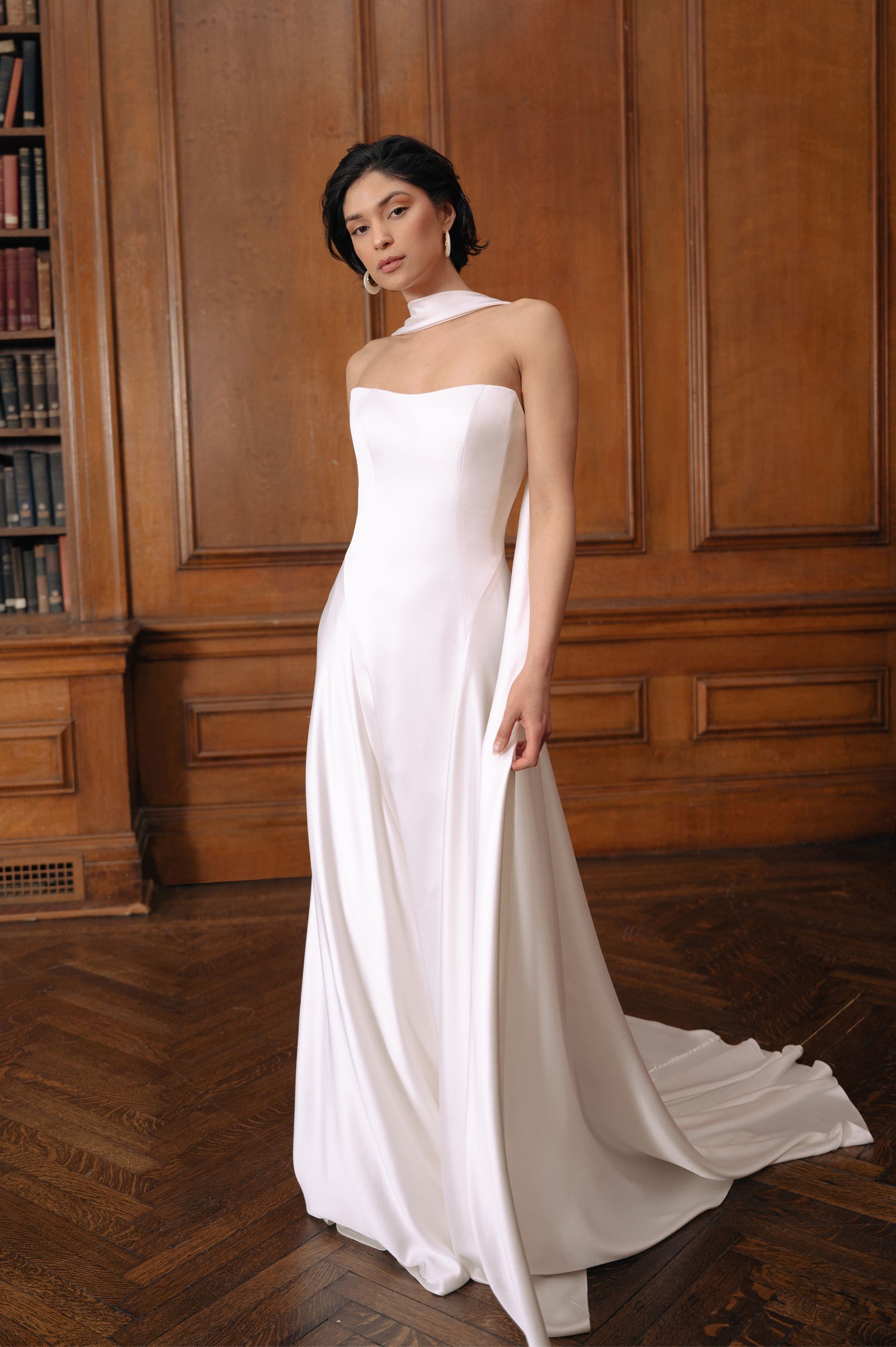 A person wears a minimalist white satin bridal gown with a halter neckline in a room with wood-paneled walls and flooring.