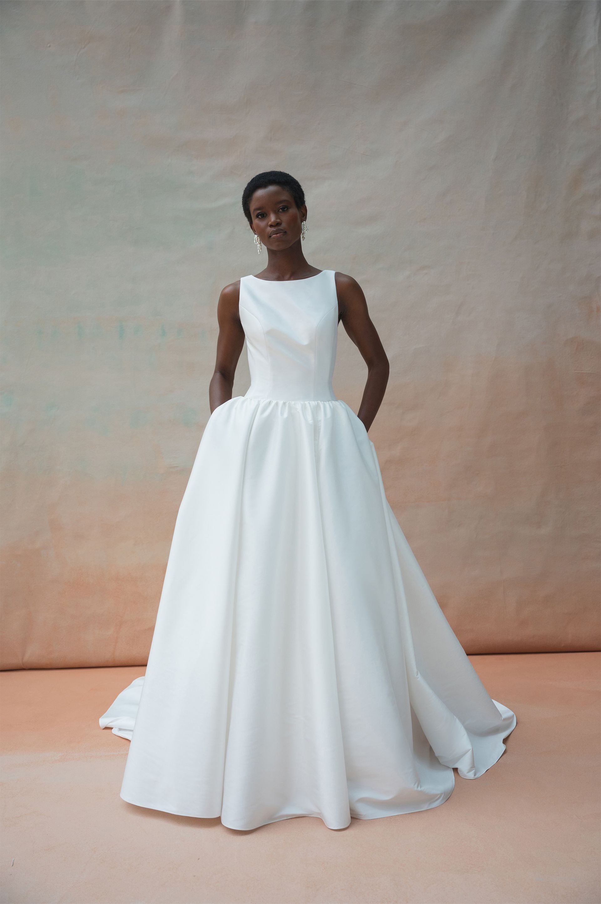A person wears a minimalist white A-line wedding dress with a high neckline and full skirt against a muted peach backdrop.