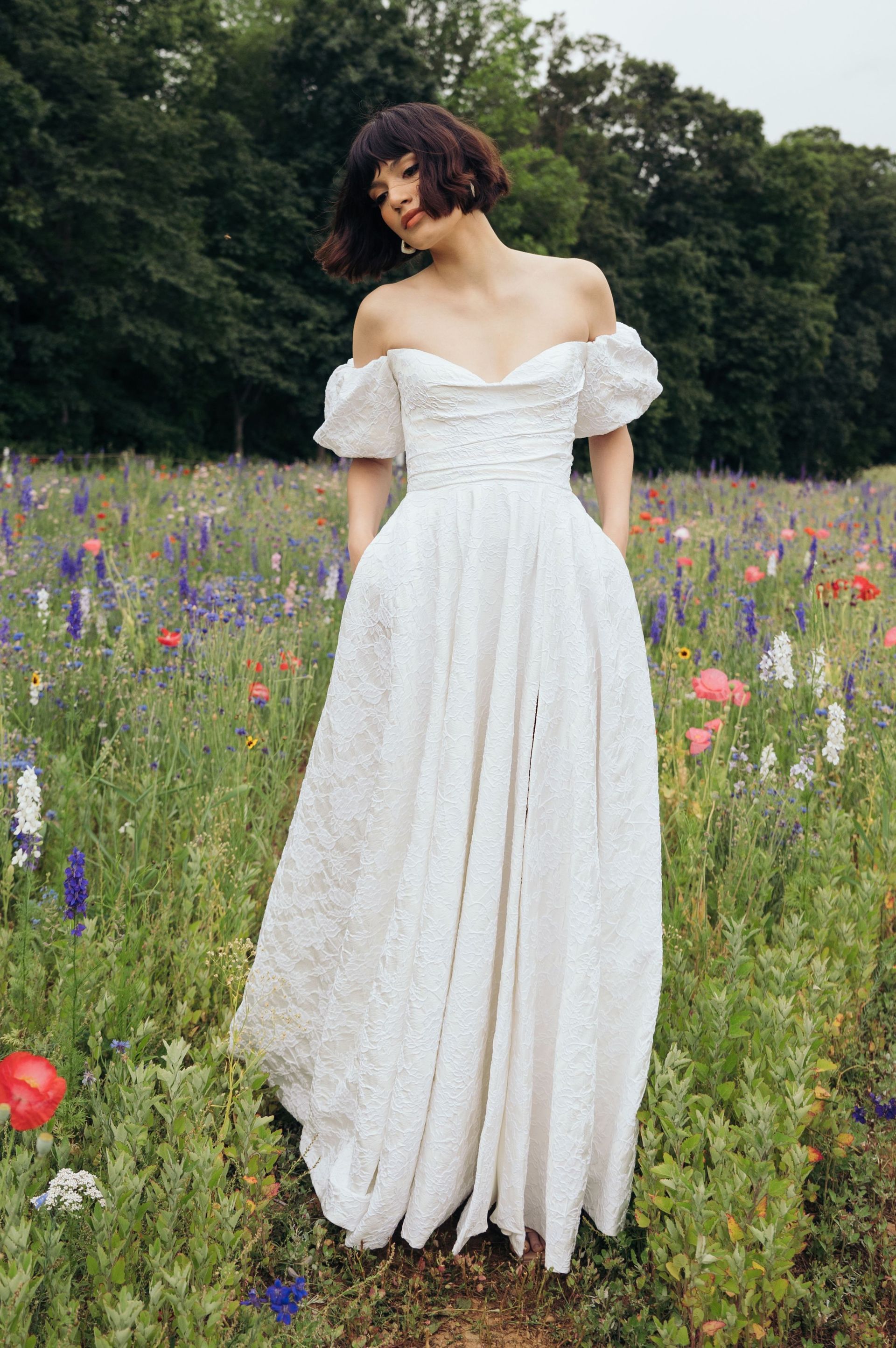 A person with short dark hair in a white, off-the-shoulder, textured gown stands in a field of wildflowers and trees.