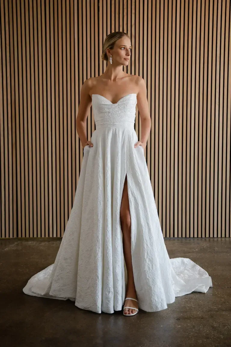 A model wears a strapless, white floral-patterned A-line wedding dress with a high leg slit and pockets against a wood wall.