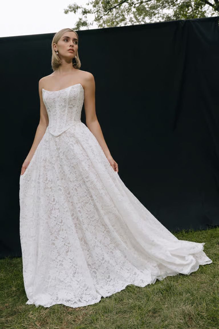 A model wearing a strapless, floor-length white lace ballgown with a structured corset bodice against a black backdrop.