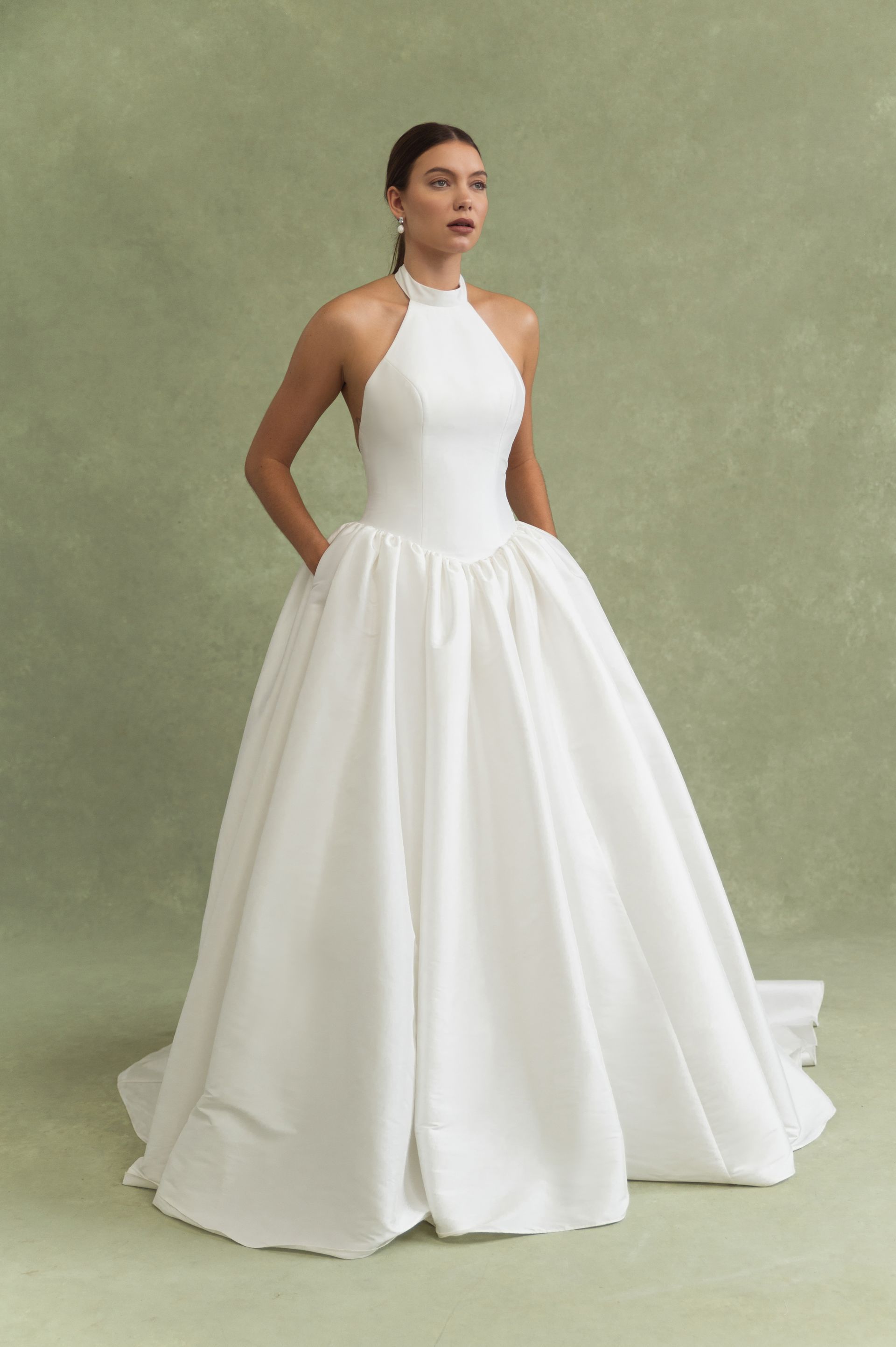 A person wears a white, floor-length ballgown with a high halter neckline and pockets against a pale green background.