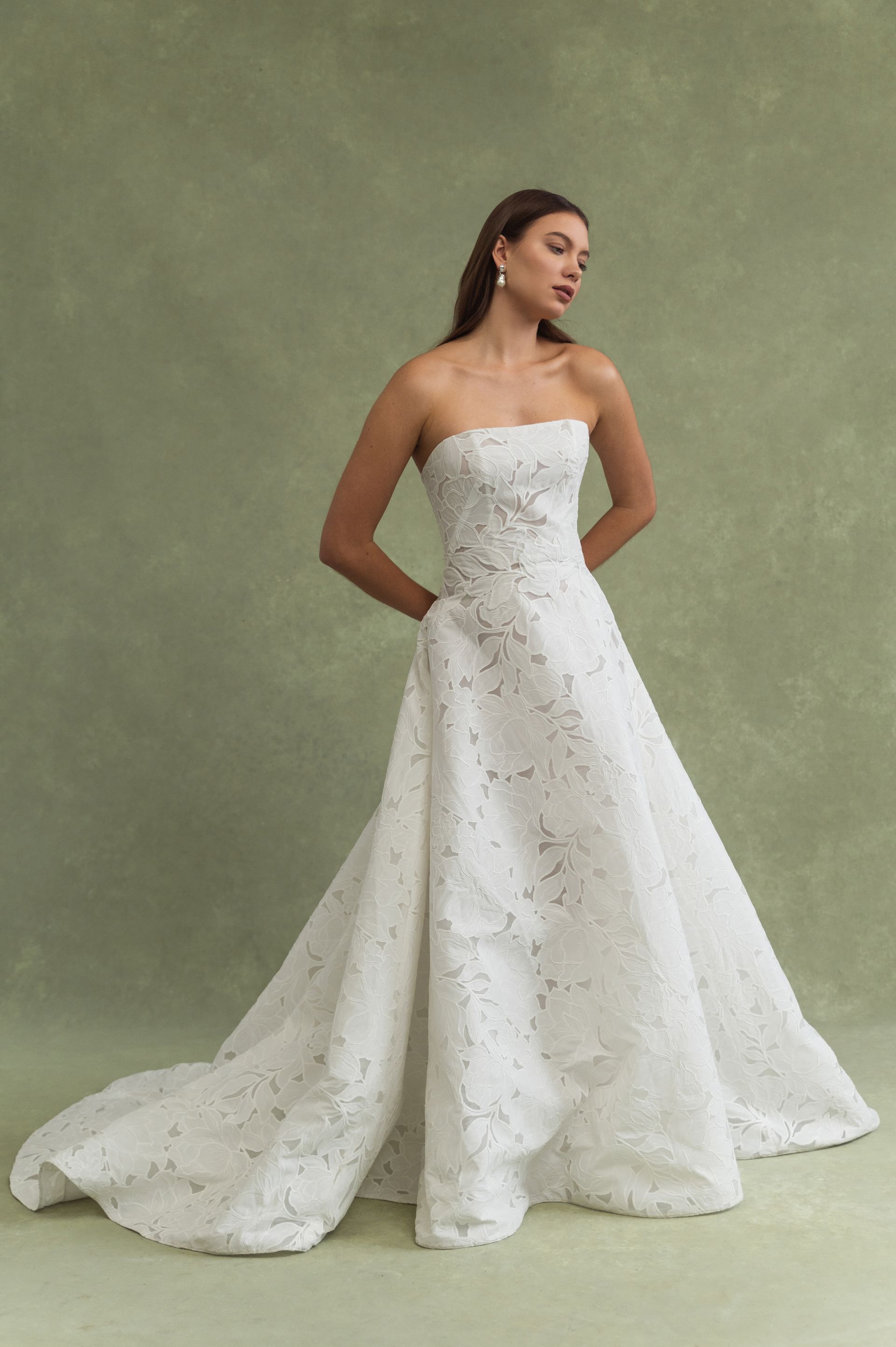 A person in a strapless, A-line white gown with delicate floral embroidery, posing against a solid green background.