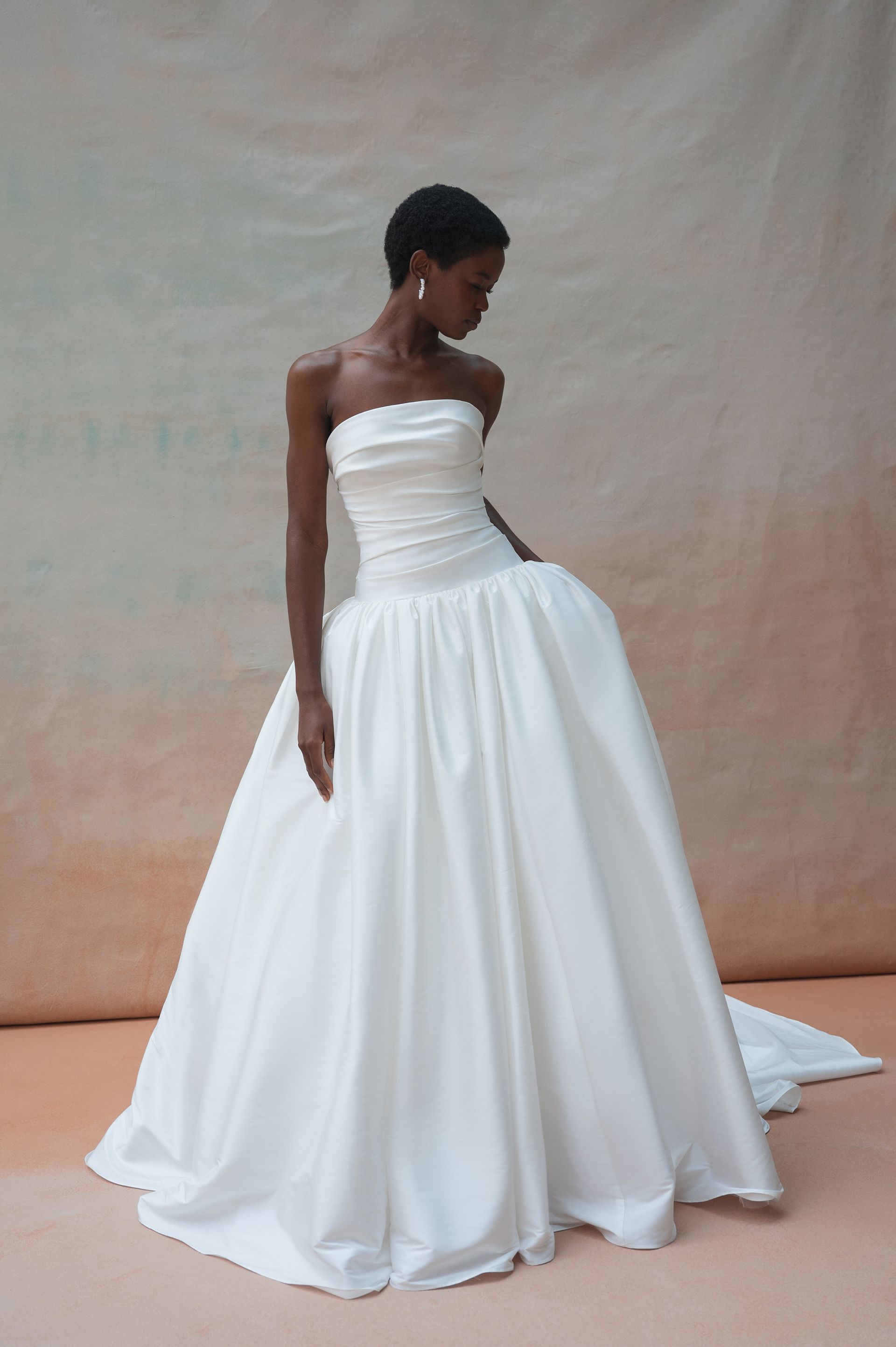 A person wears a strapless, white, full-skirted gown against a neutral-toned, draped fabric backdrop.
