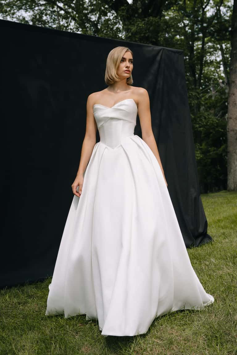 A person in a strapless, white A-line wedding gown with a fitted bodice, standing on grass before a dark backdrop.
