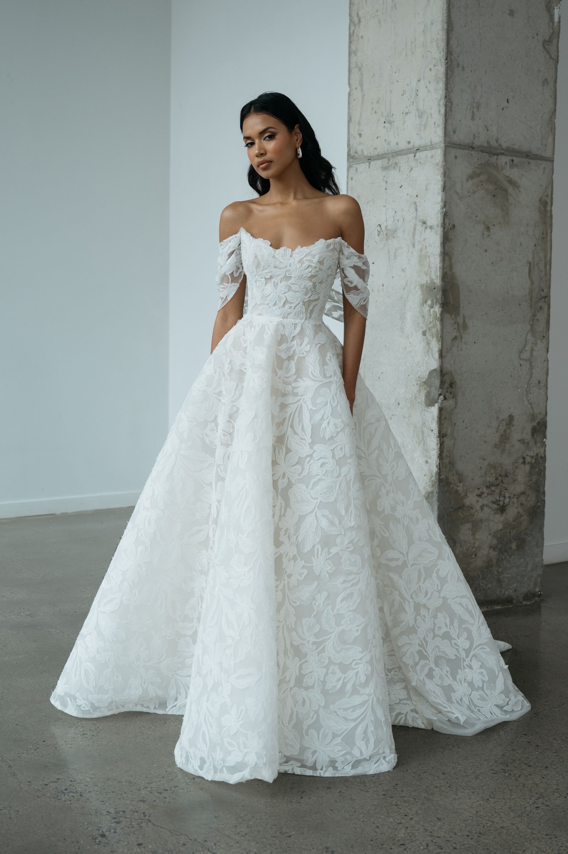 A model wearing an off-the-shoulder, white, floor-length lace wedding dress with a full skirt in a minimalist room.