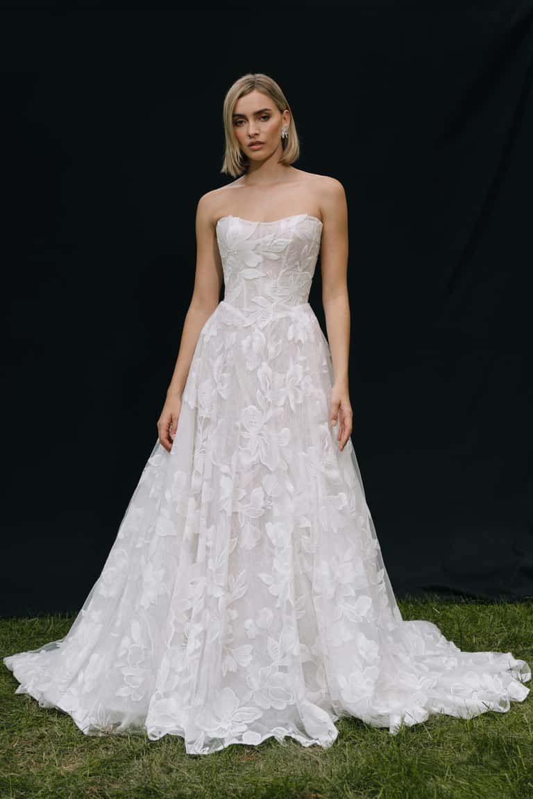 A person wearing a strapless white wedding gown with a full, textured floral skirt, standing against a black backdrop.