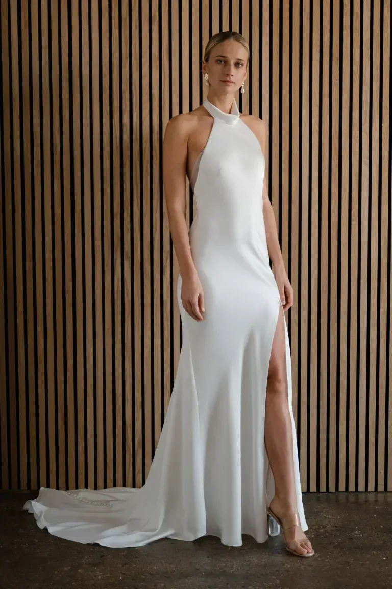 A person wearing a floor-length white satin halter wedding dress with a high leg slit, posing against a slatted wall.