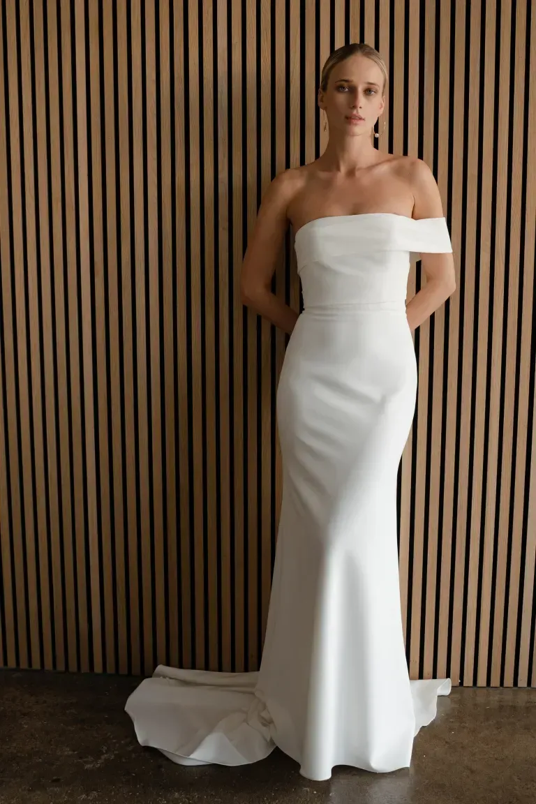 A model wearing a white, strapless, floor-length crepe wedding gown with a draped off-the-shoulder detail and a small train.