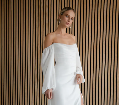 A model wearing a white off-the-shoulder wedding dress with long, draped sleeves, posed against a vertical wood-slat wall.