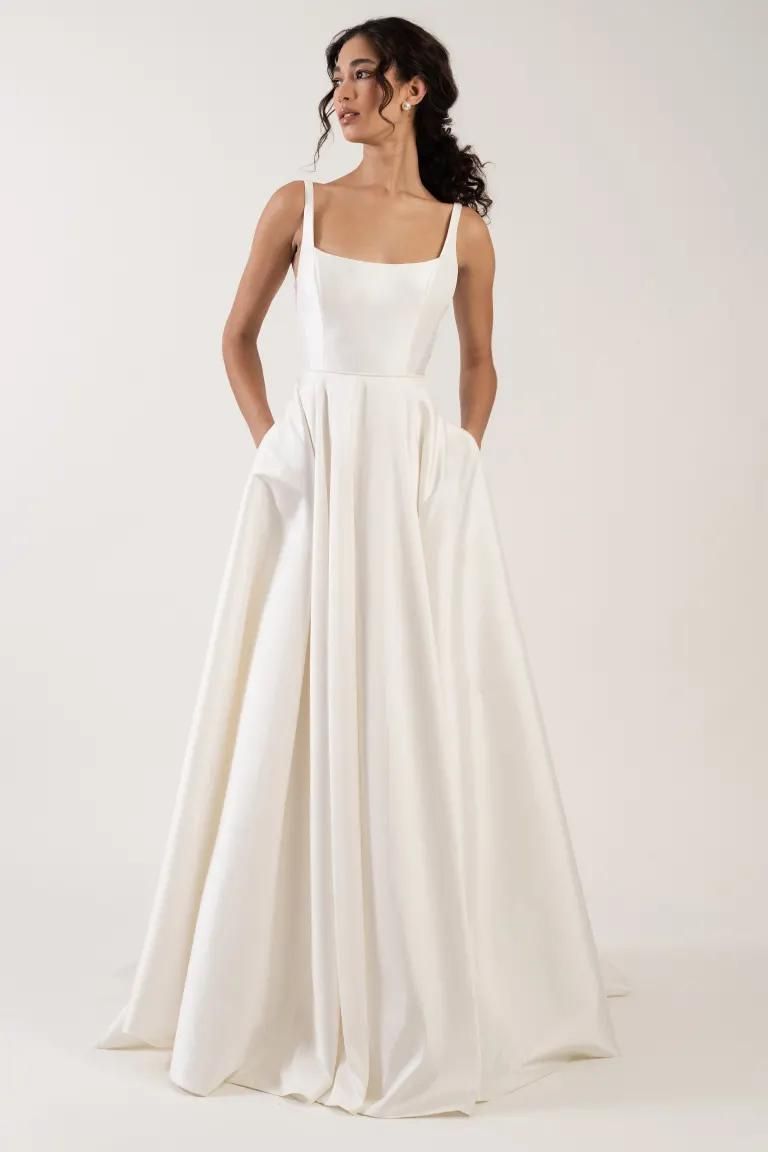 A person modeling a white, floor-length A-line wedding dress with a square neckline, spaghetti straps, and pockets.