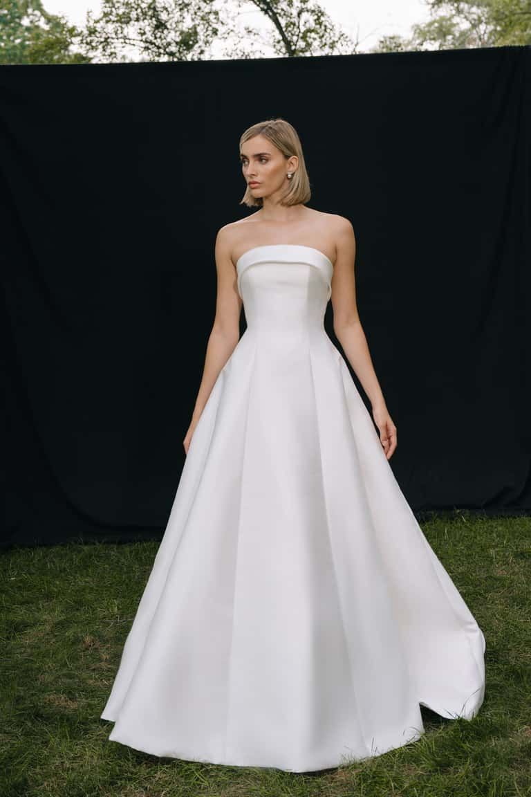 A model wearing a strapless, floor-length white satin A-line gown, standing on grass against a black backdrop.