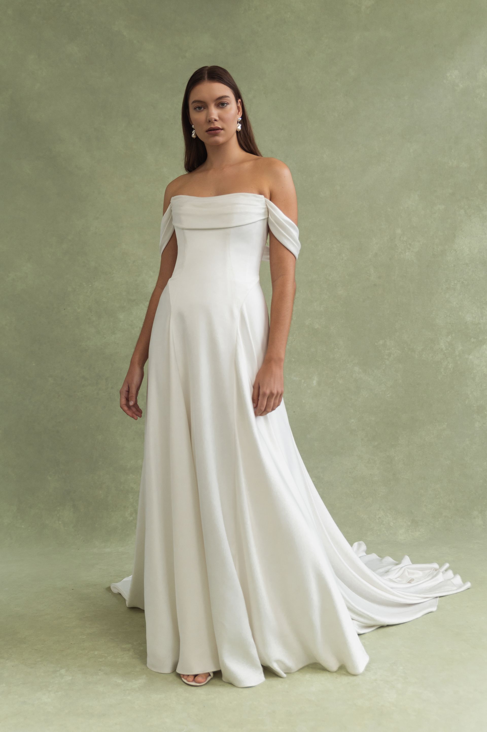 A person wears a minimalist, floor-length off-the-shoulder white wedding gown against a sage green textured background.