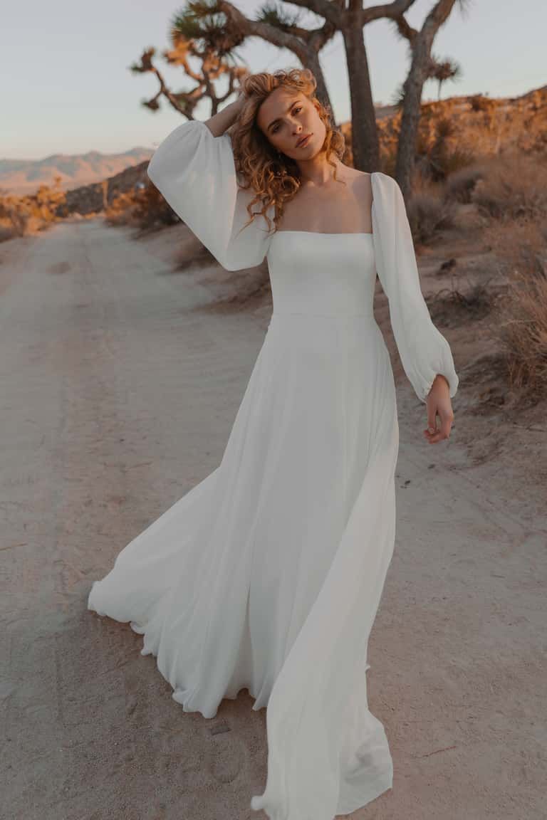 A person with wavy hair walks on a dirt path in a desert, wearing a long-sleeved, floor-length white dress at sunset.