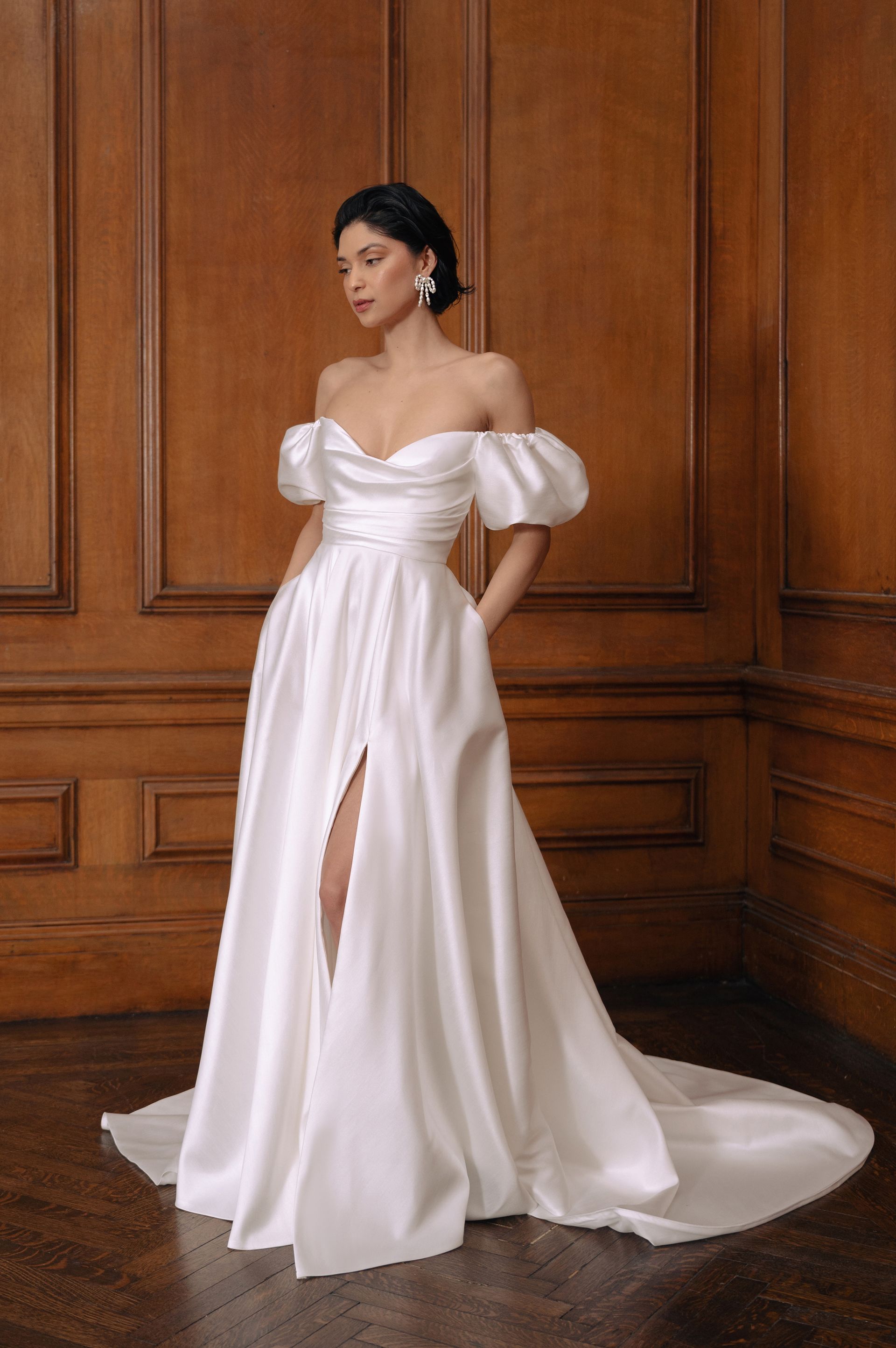 A model wearing a white satin off-the-shoulder wedding dress with puff sleeves, a sweetheart neckline, and a leg slit.