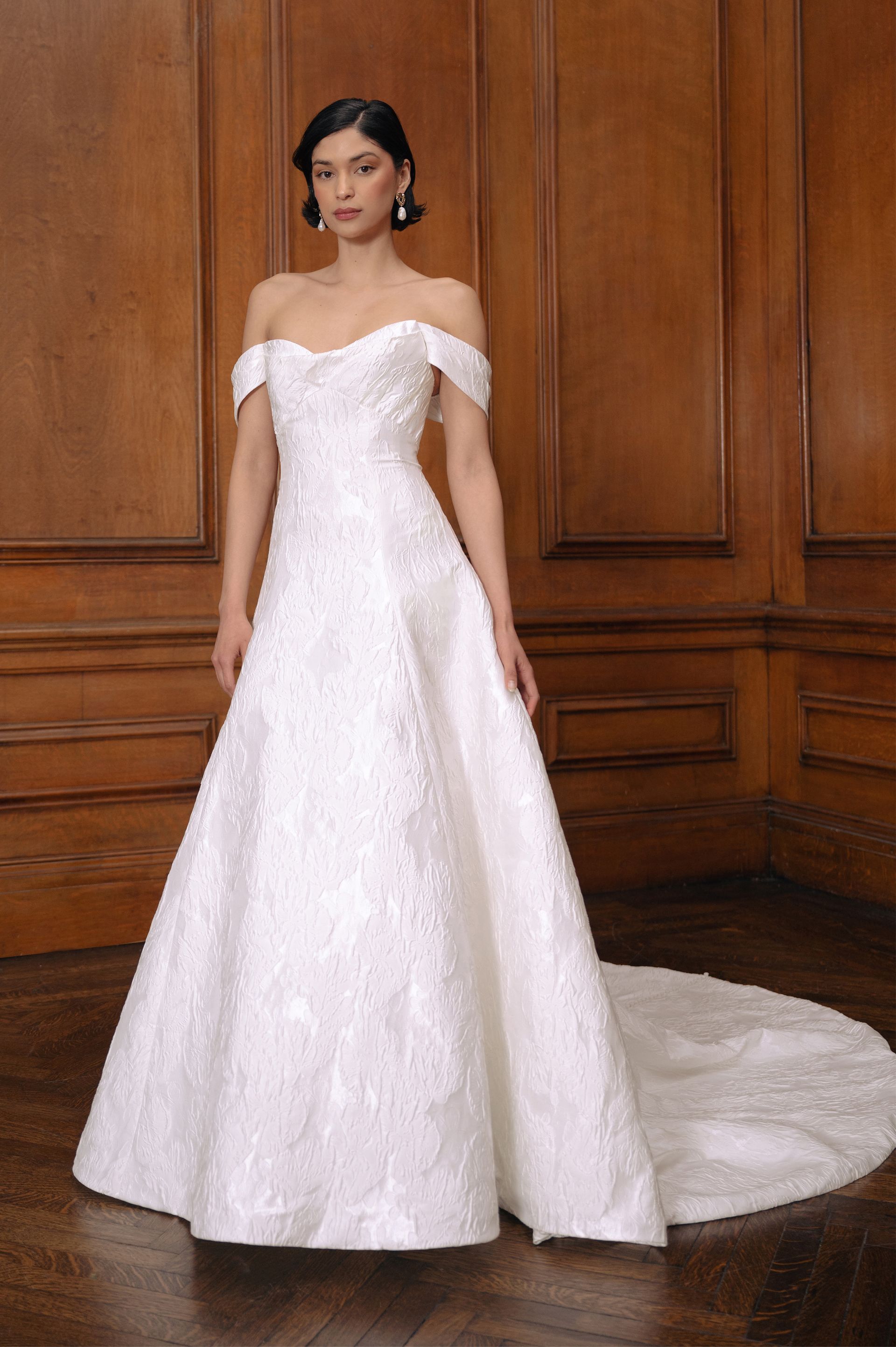 A model wears an off-the-shoulder, white, textured A-line wedding dress with a train in a room with wood-paneled walls.