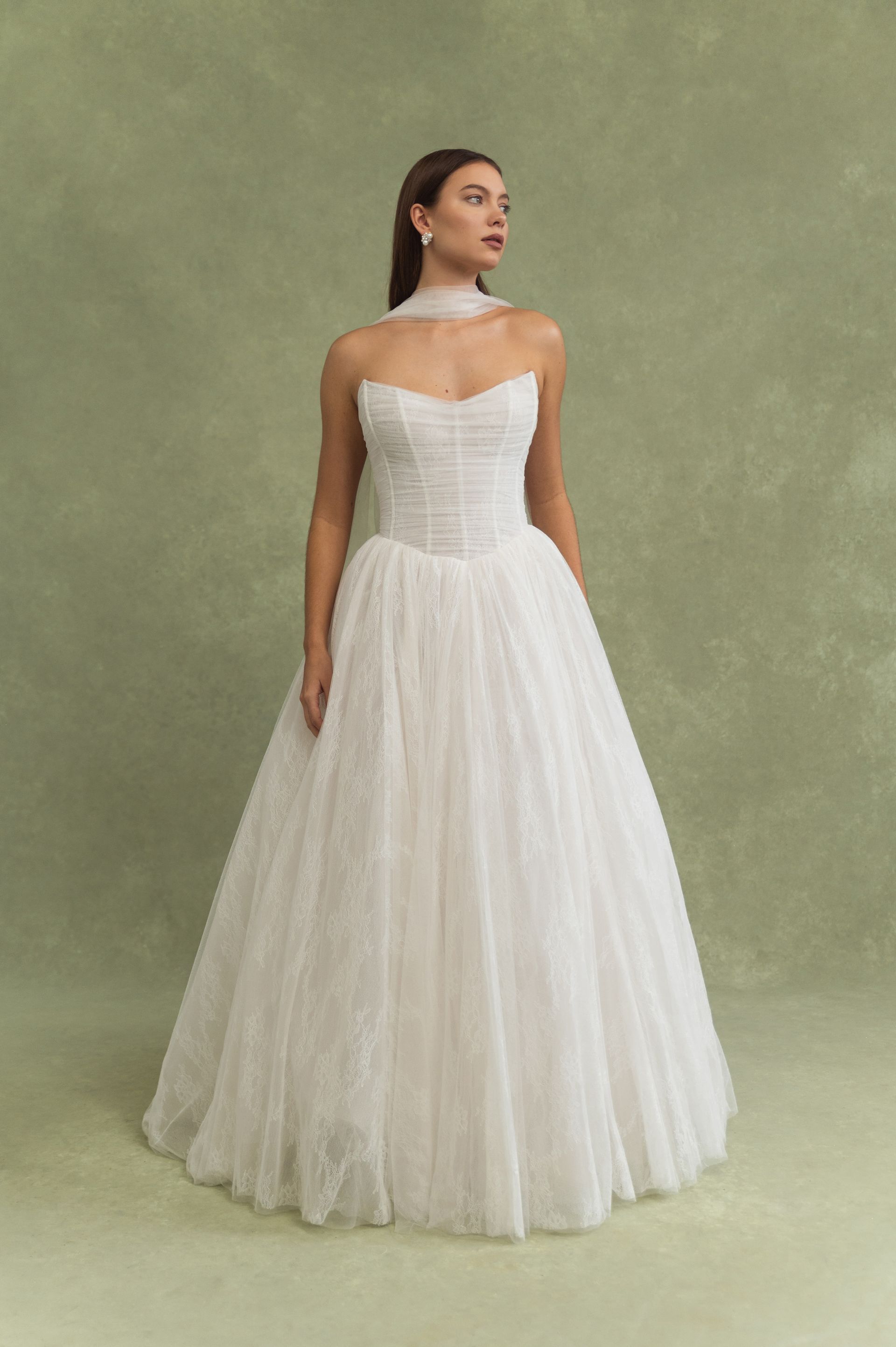 A model wears a white, strapless, floor-length tulle wedding gown with a matching sheer neck scarf against a green backdrop.