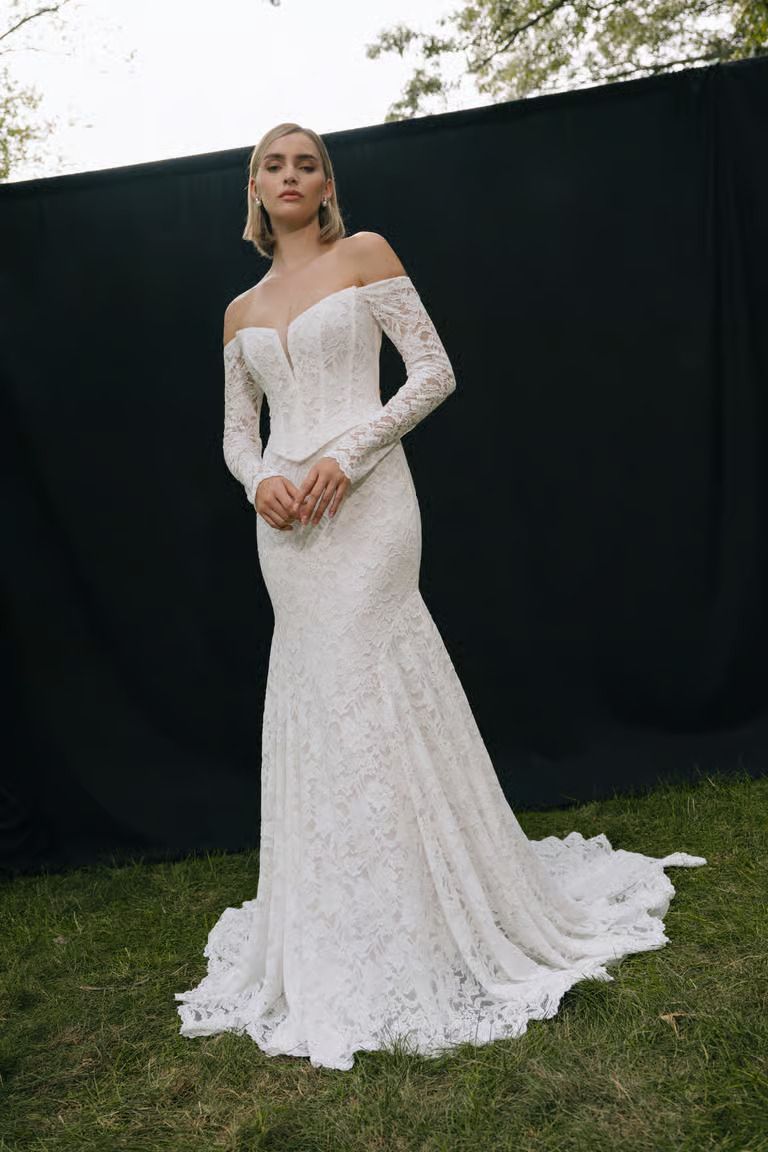 A person wearing a long-sleeved, off-the-shoulder white lace wedding dress with a train, standing on grass.