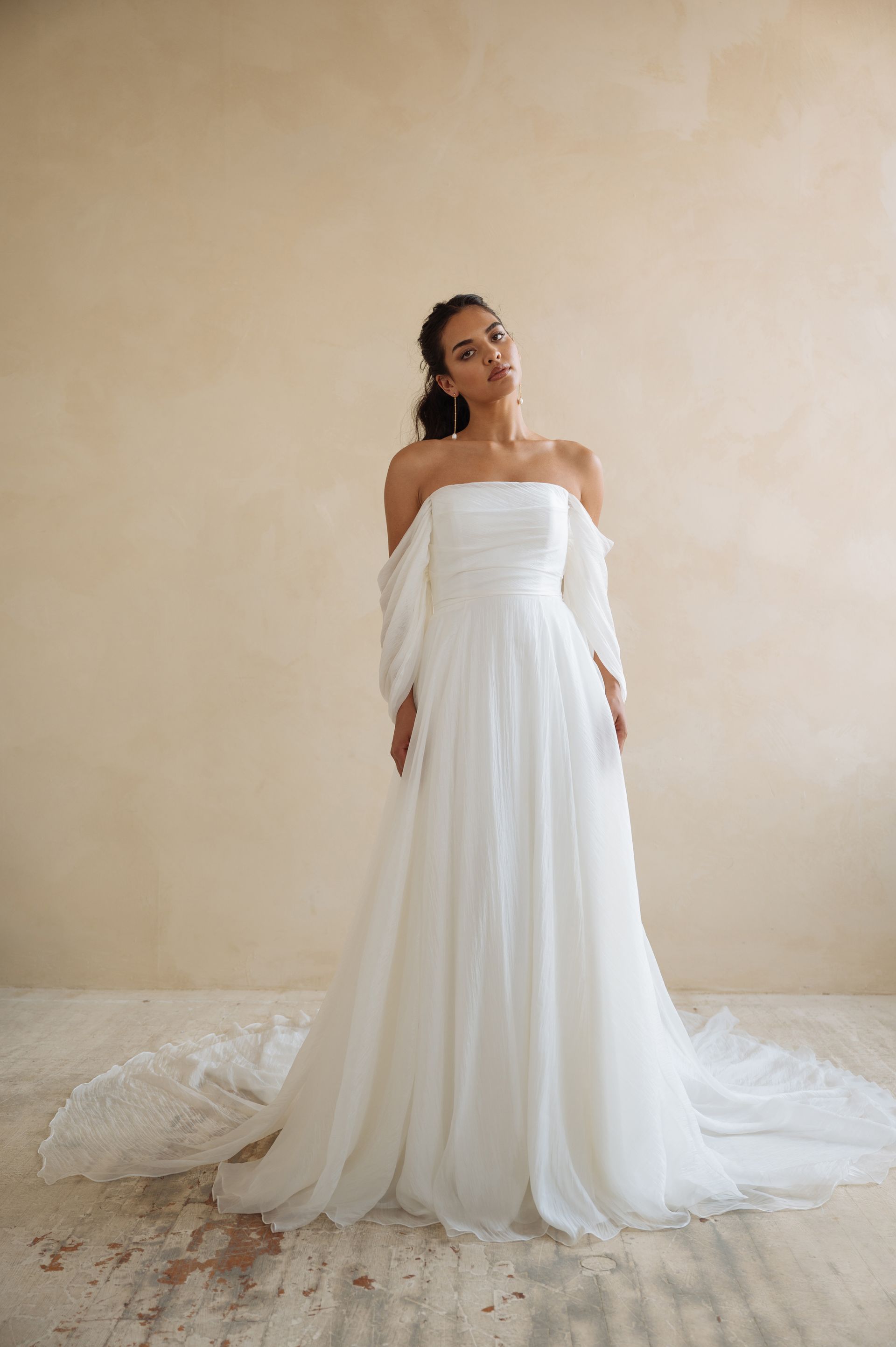 A person wearing a white, off-the-shoulder A-line wedding dress with a train, standing against a neutral textured wall.