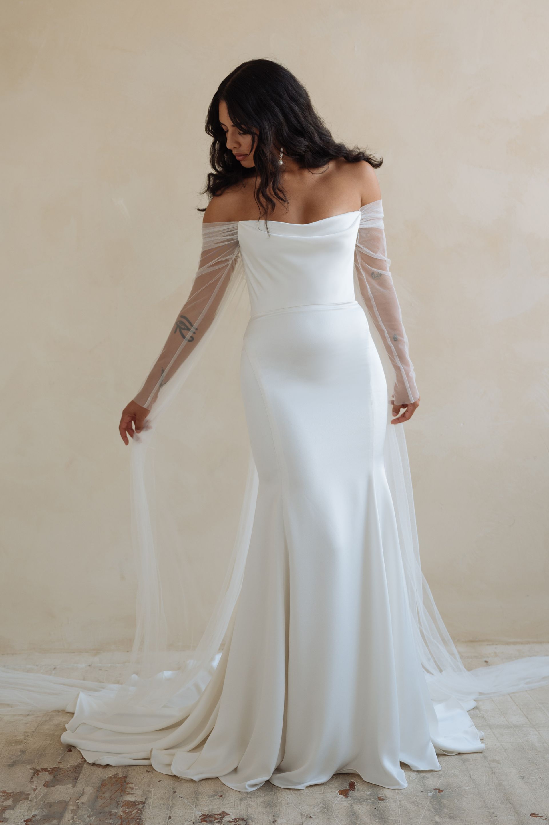 A person models an off-the-shoulder, white floor-length wedding gown with sheer, beaded long sleeves and a long train.