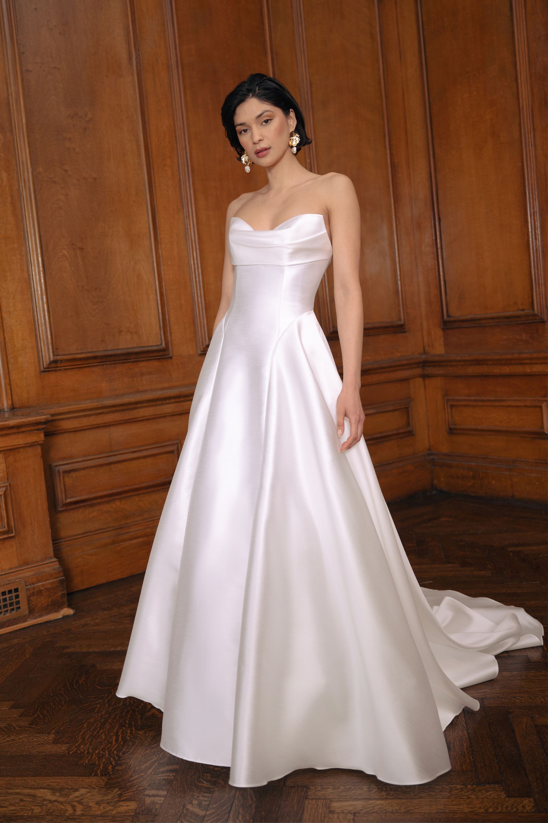 A person wearing a white, strapless, A-line satin wedding gown with a train in a room with wood-paneled walls.
