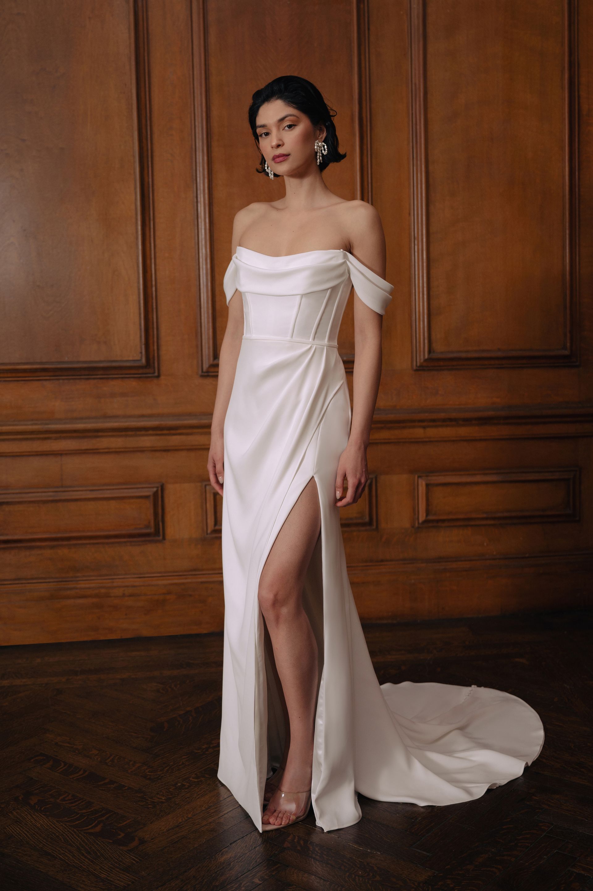 A model wearing a white, off-the-shoulder, satin floor-length wedding gown with a high slit and train in a wood-paneled room.
