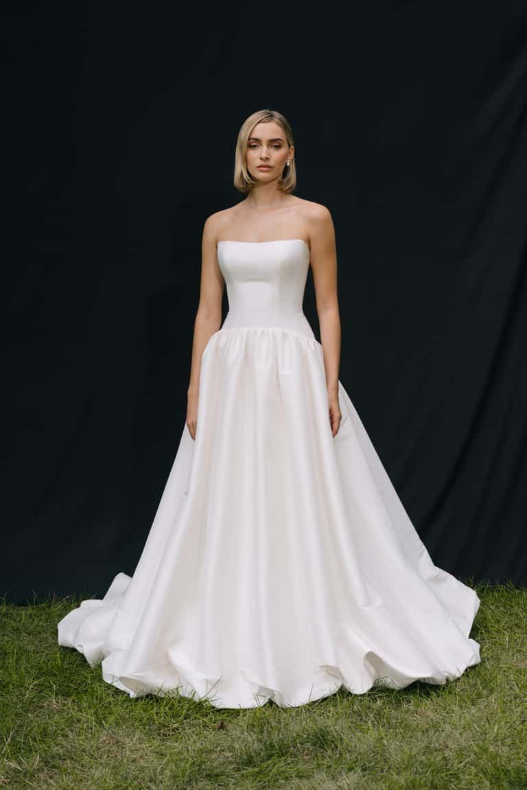 A person wears a strapless, white A-line gown with a fitted bodice and full skirt, standing on grass