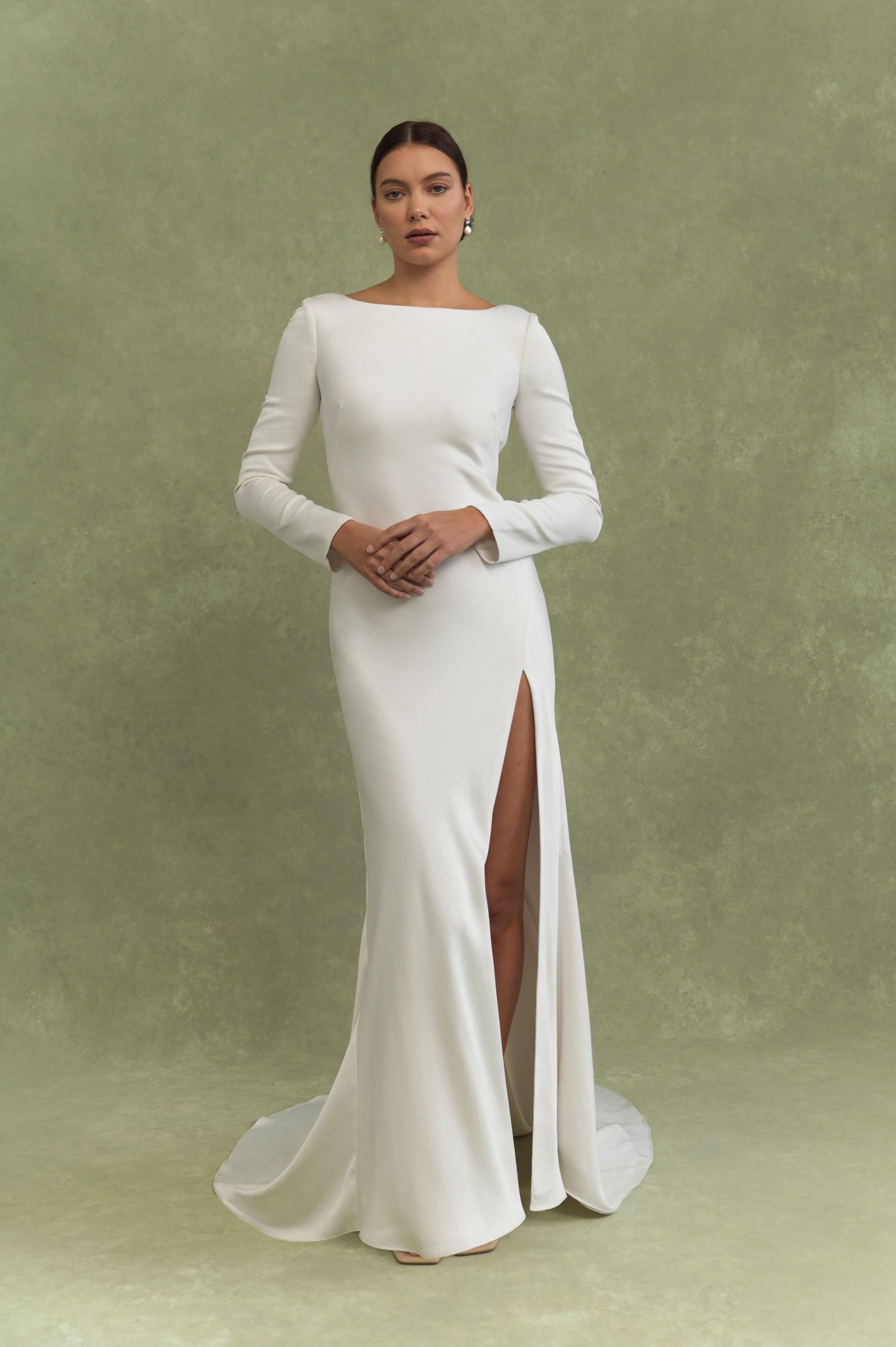A long-sleeved, white floor-length gown with a high side slit, worn by a person against a solid green background.