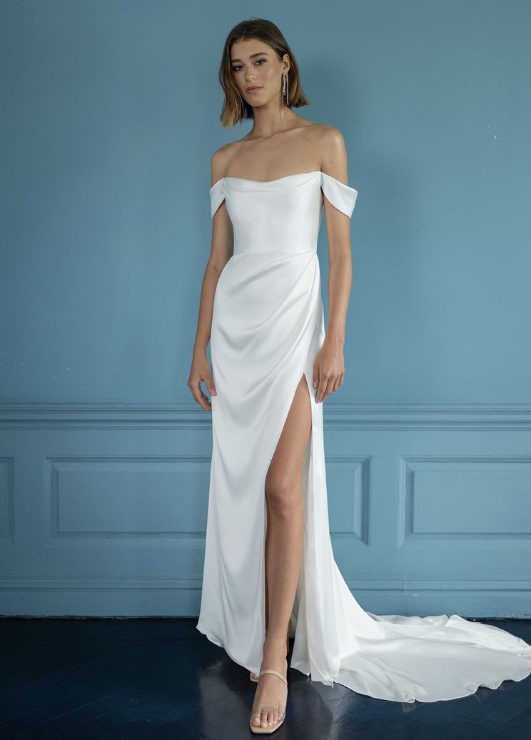 A person in a white off-the-shoulder, floor-length silk dress with a high side slit stands against a blue wall.