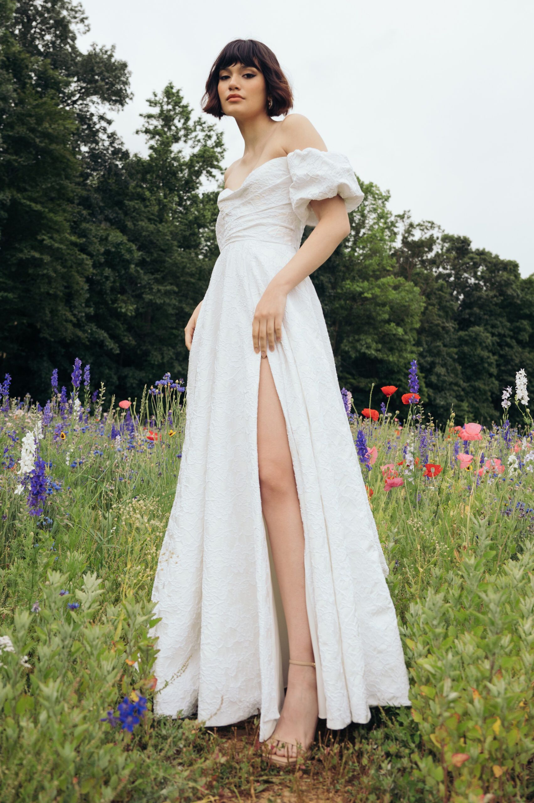 A person in a white, off-the-shoulder, floor-length dress with a high side slit stands in a meadow of wildflowers.