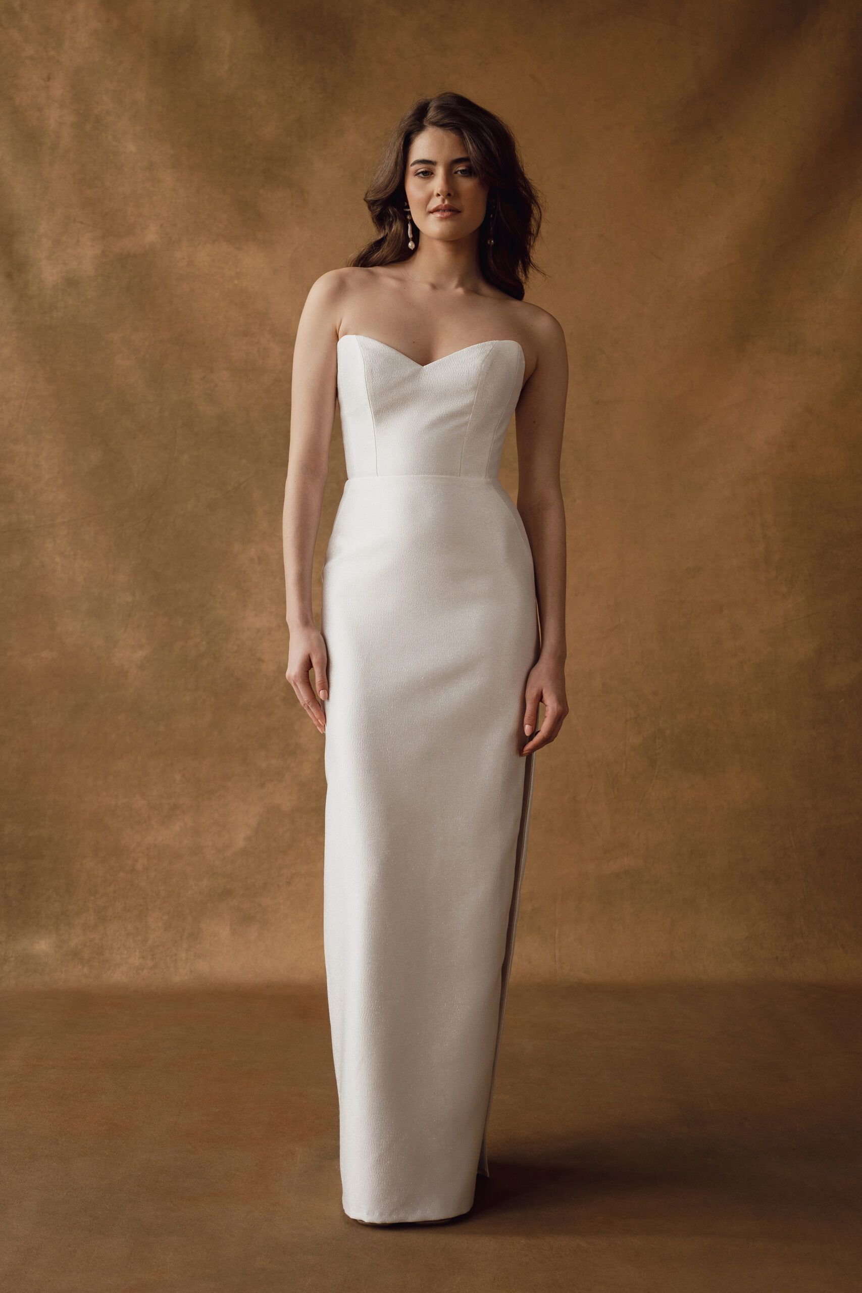 A person poses in a strapless, floor-length white wedding dress against a textured, warm-toned brown background.