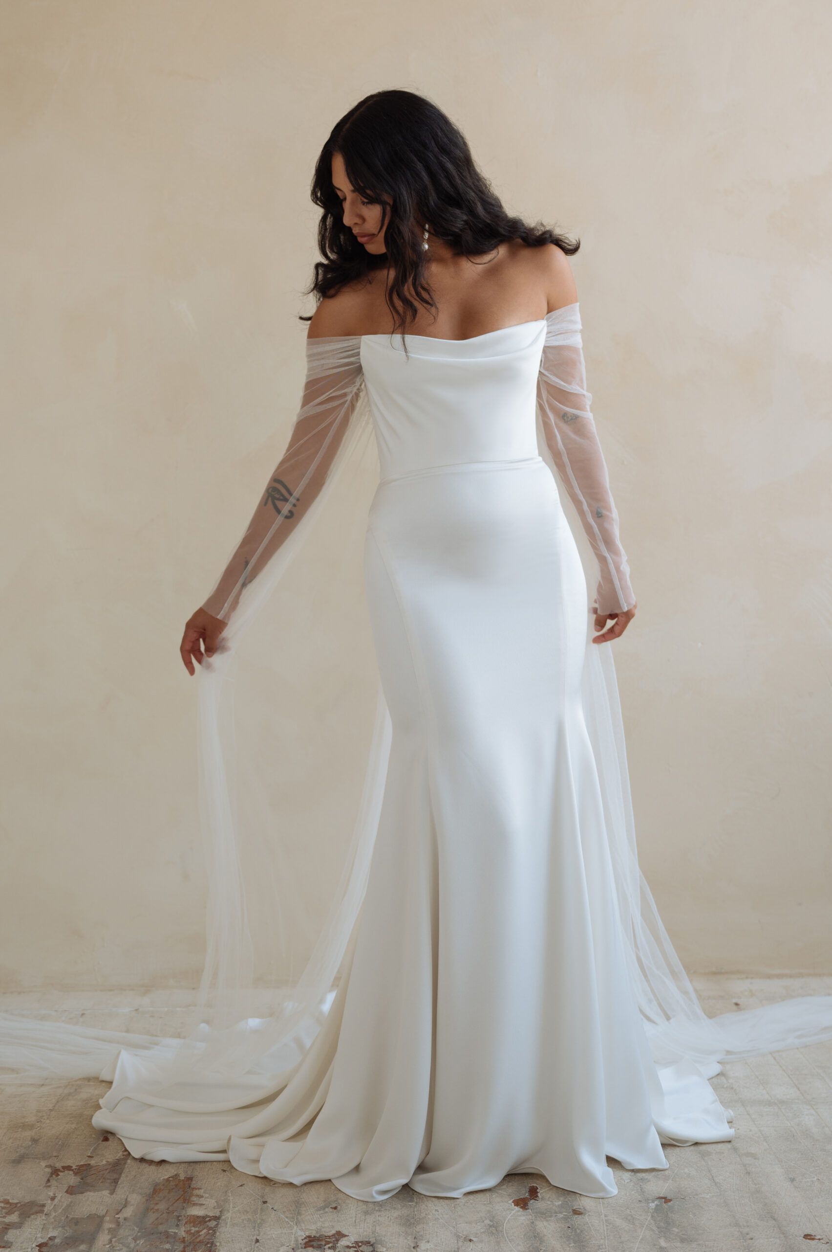 A woman in an off-the-shoulder, white floor-length wedding gown with sheer, beaded, floor-length sleeves.