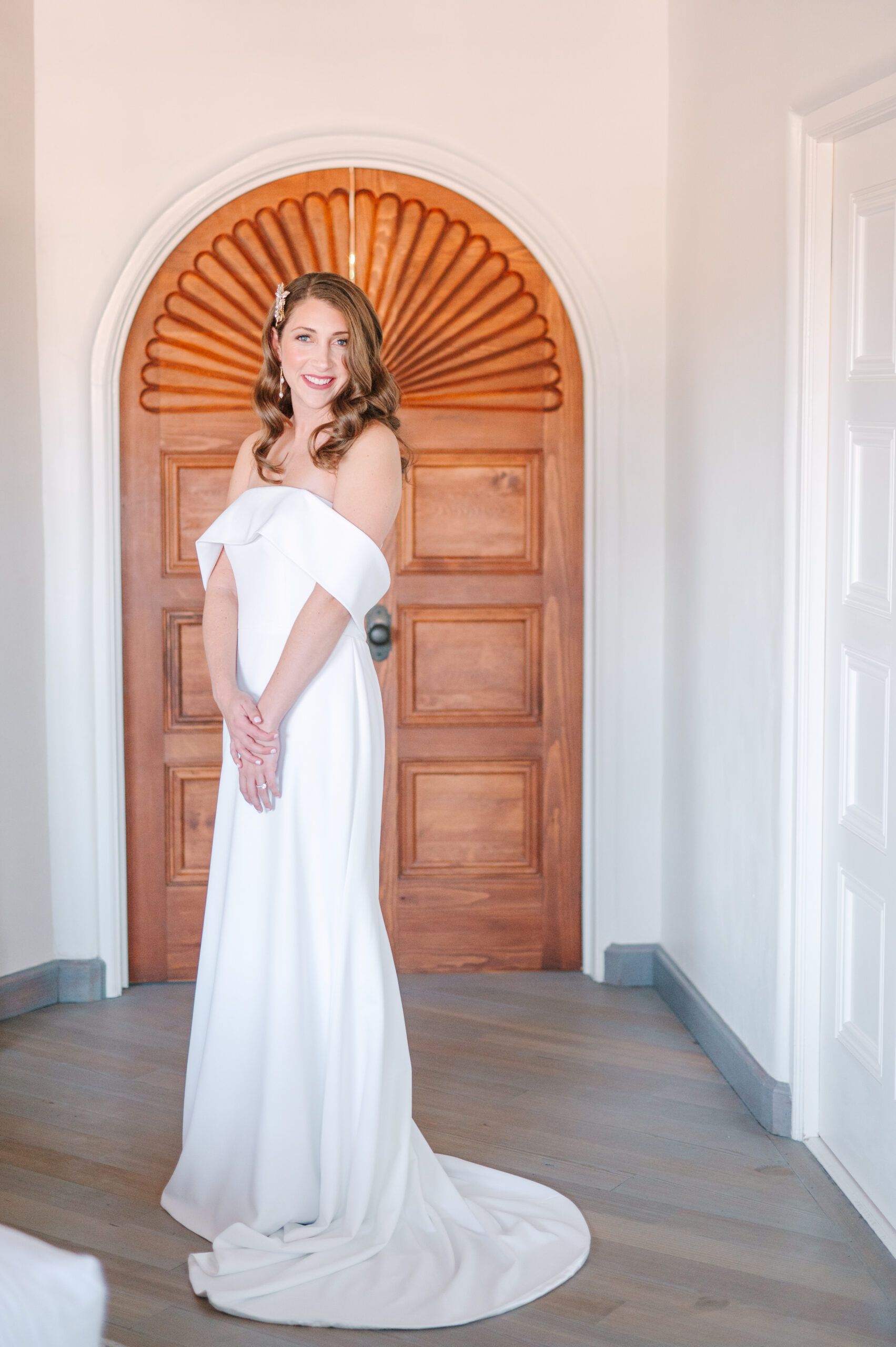 A smiling person wearing a white off-the-shoulder wedding dress stands in front of a decorative wooden arched door.
