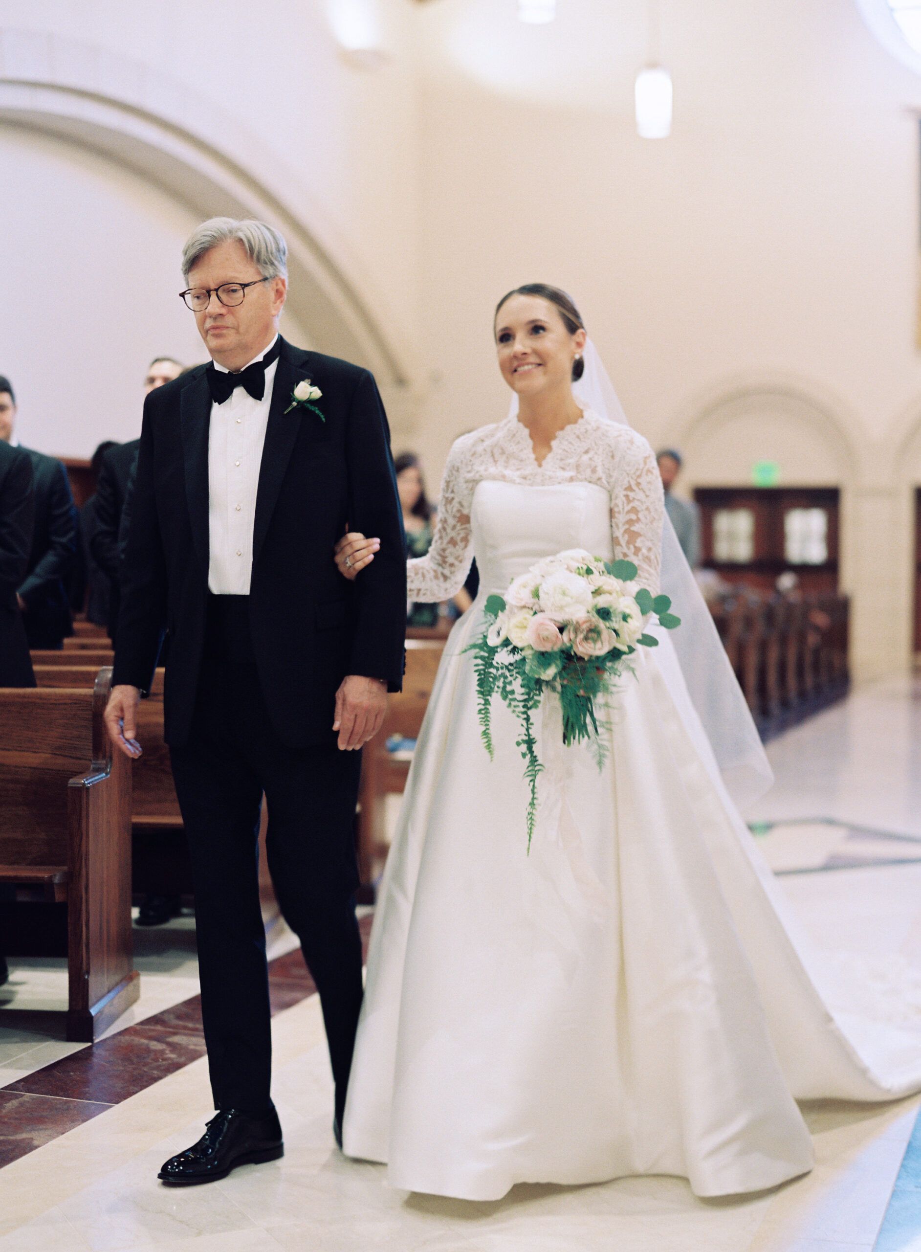 A person in a white wedding gown holding a bouquet walks down a church aisle on the arm of a person in a tuxedo.