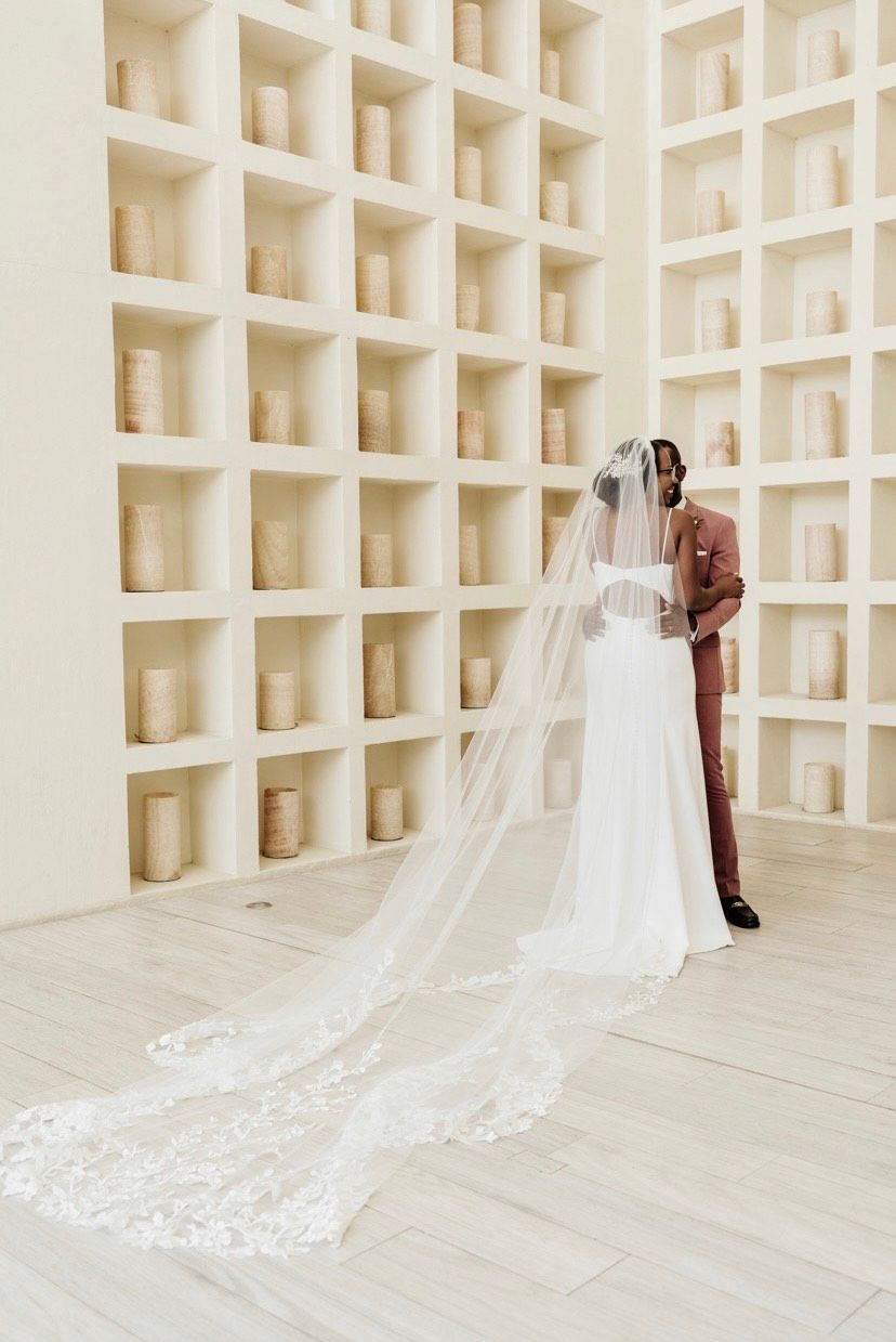 A couple embraces in front of a grid of lit candles in an off-white stone room. The bride wears a long, lace-trimmed veil.