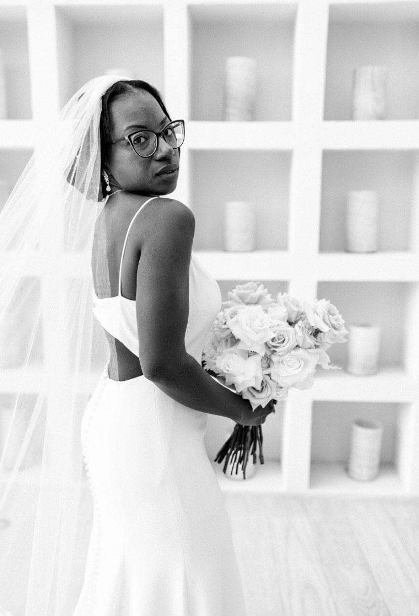 A person in a wedding dress and veil holding a bouquet, looking over their shoulder in front of white cubby shelves.
