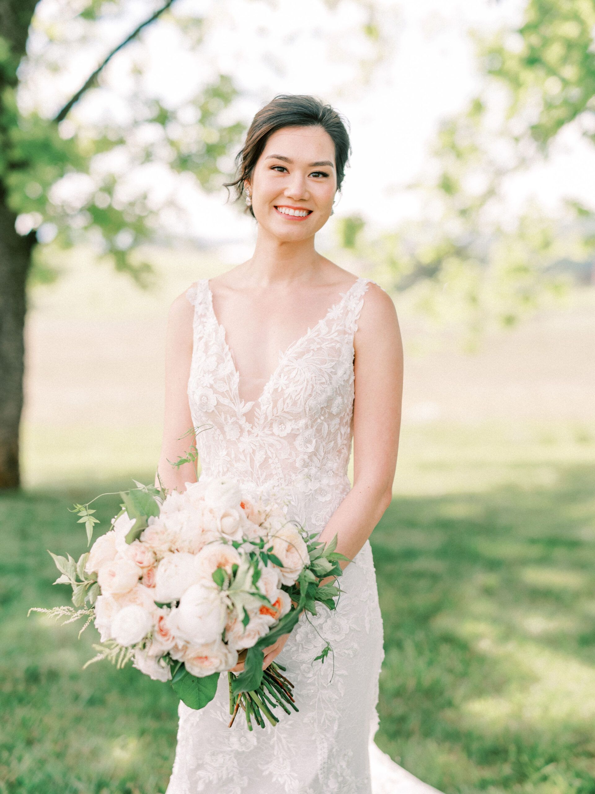 A smiling person in a lace wedding dress holds a floral bouquet outdoors in a grassy field.