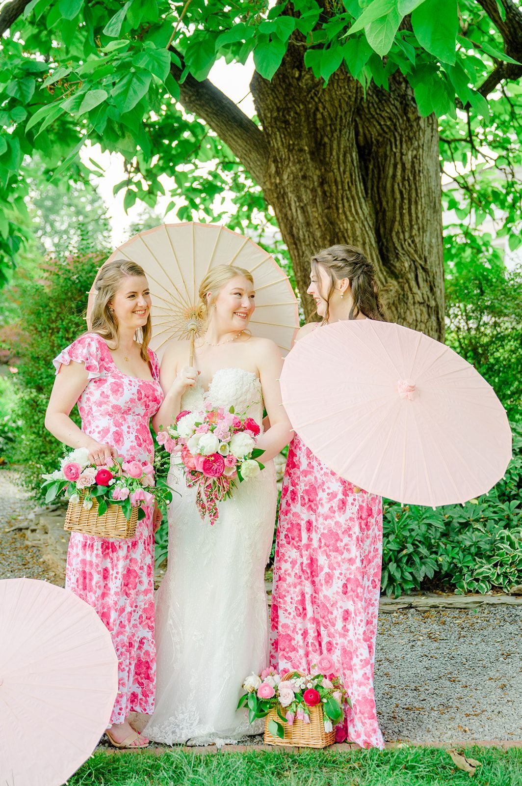 A bride in a white gown and two people in pink floral dresses hold paper parasols and flower baskets under a large tree.
