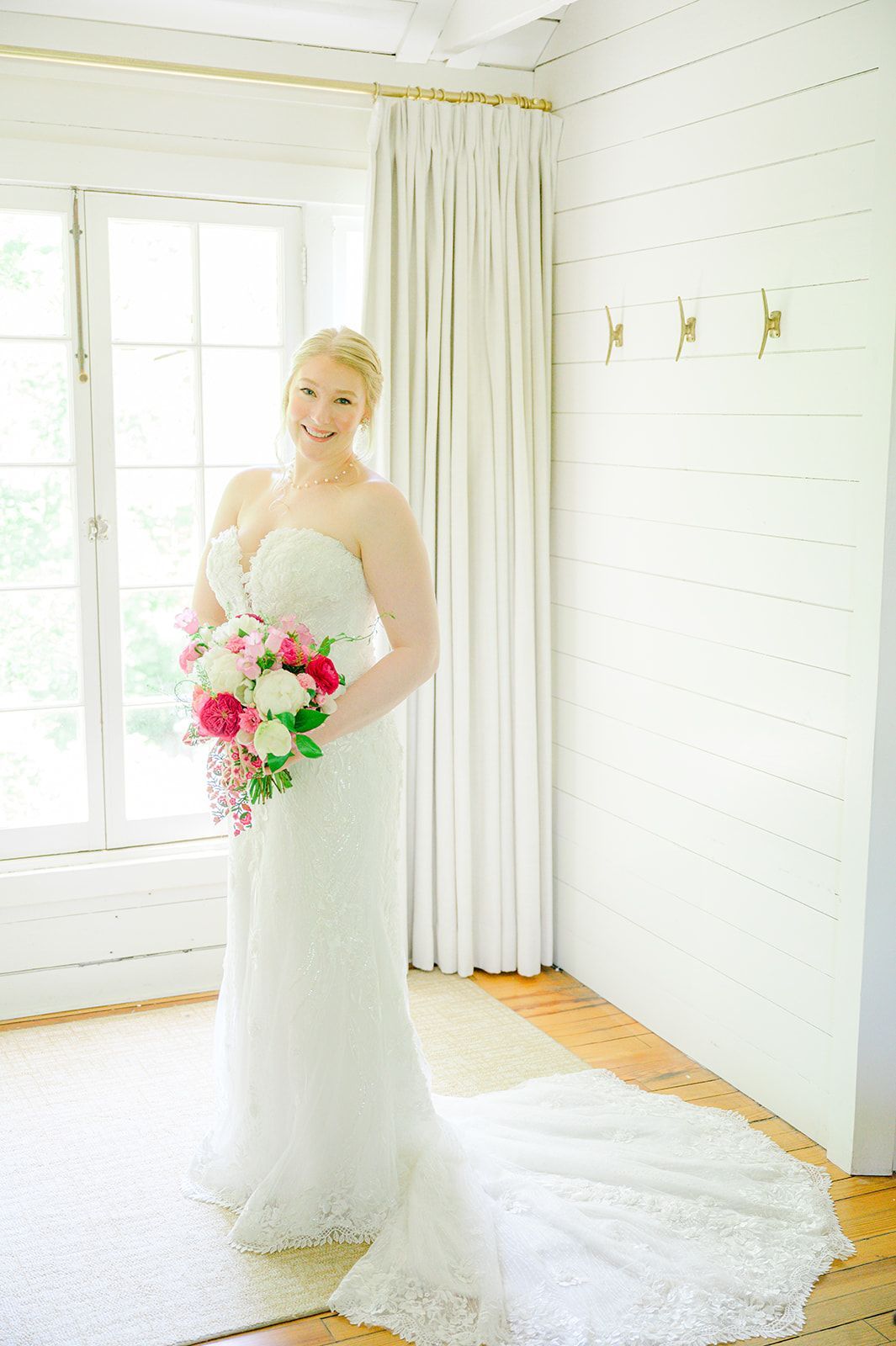 A person in a white wedding gown holding a vibrant bouquet of pink and white flowers stands in a bright, rustic room.