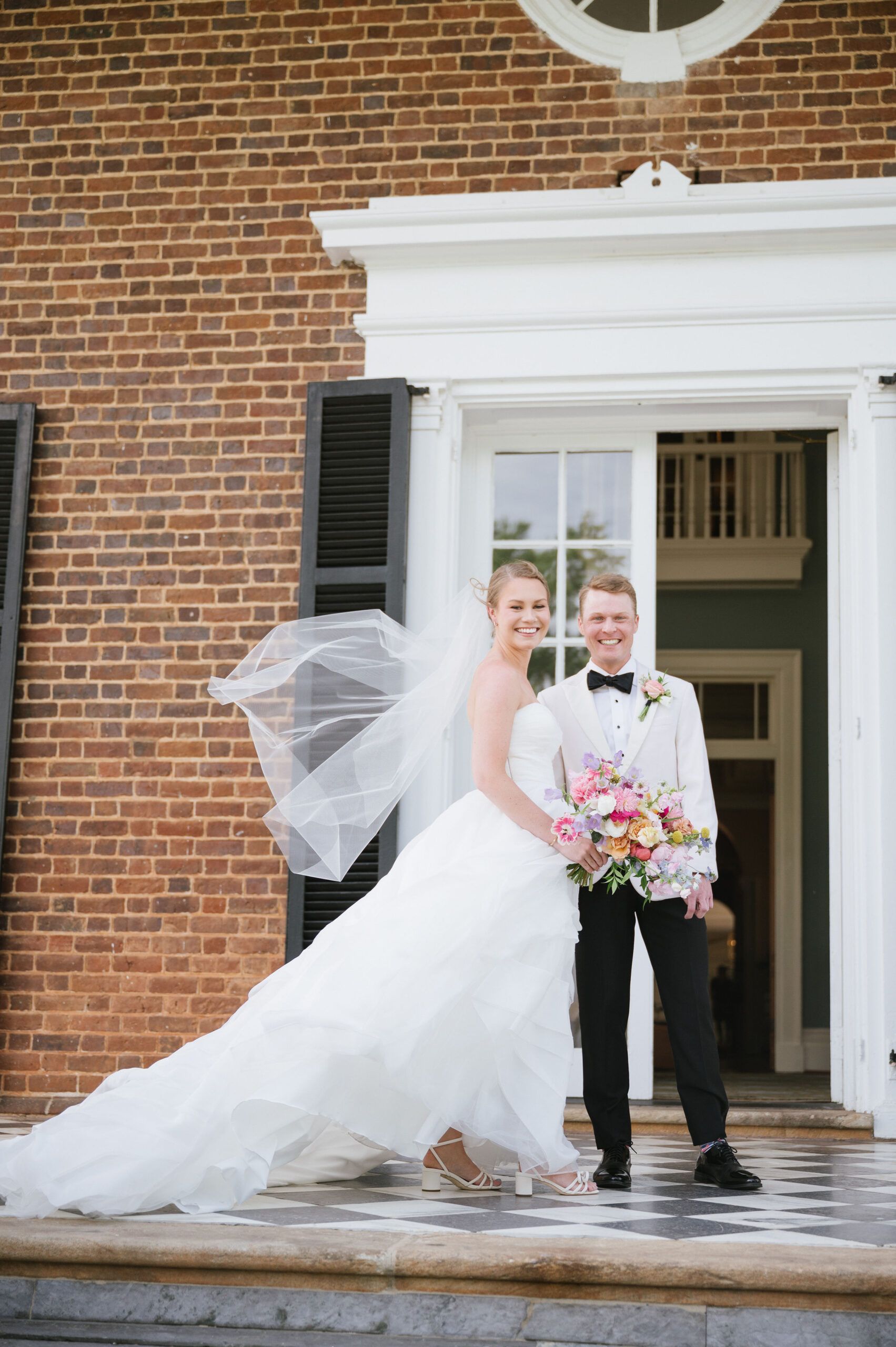 A couple stands in front of a brick building, the bride in a flowing wedding dress with a veil and the groom in a tuxedo.