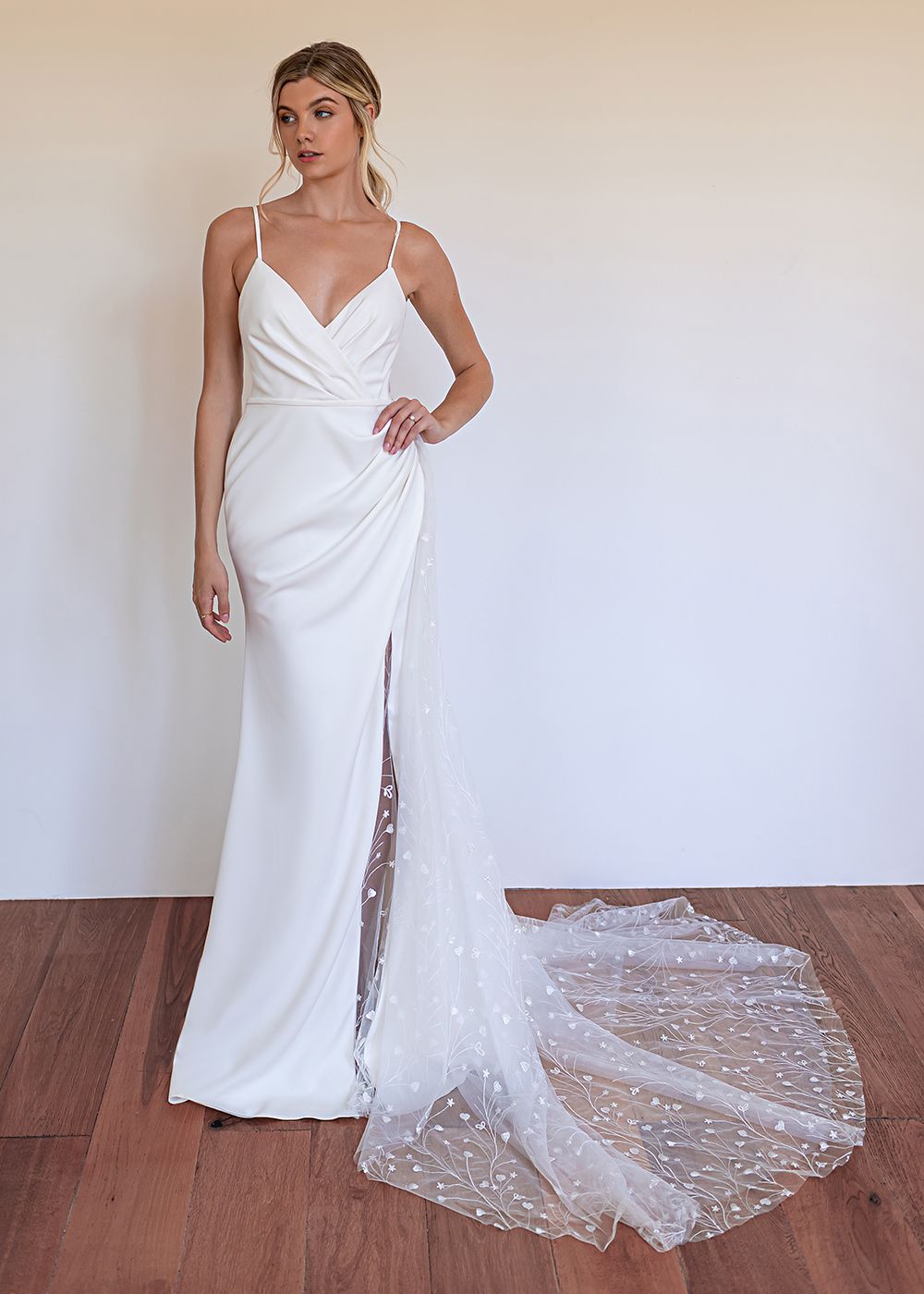 A person modeling a white, sleeveless, floor-length wedding gown with a thigh-high slit and a sparkling tulle train.