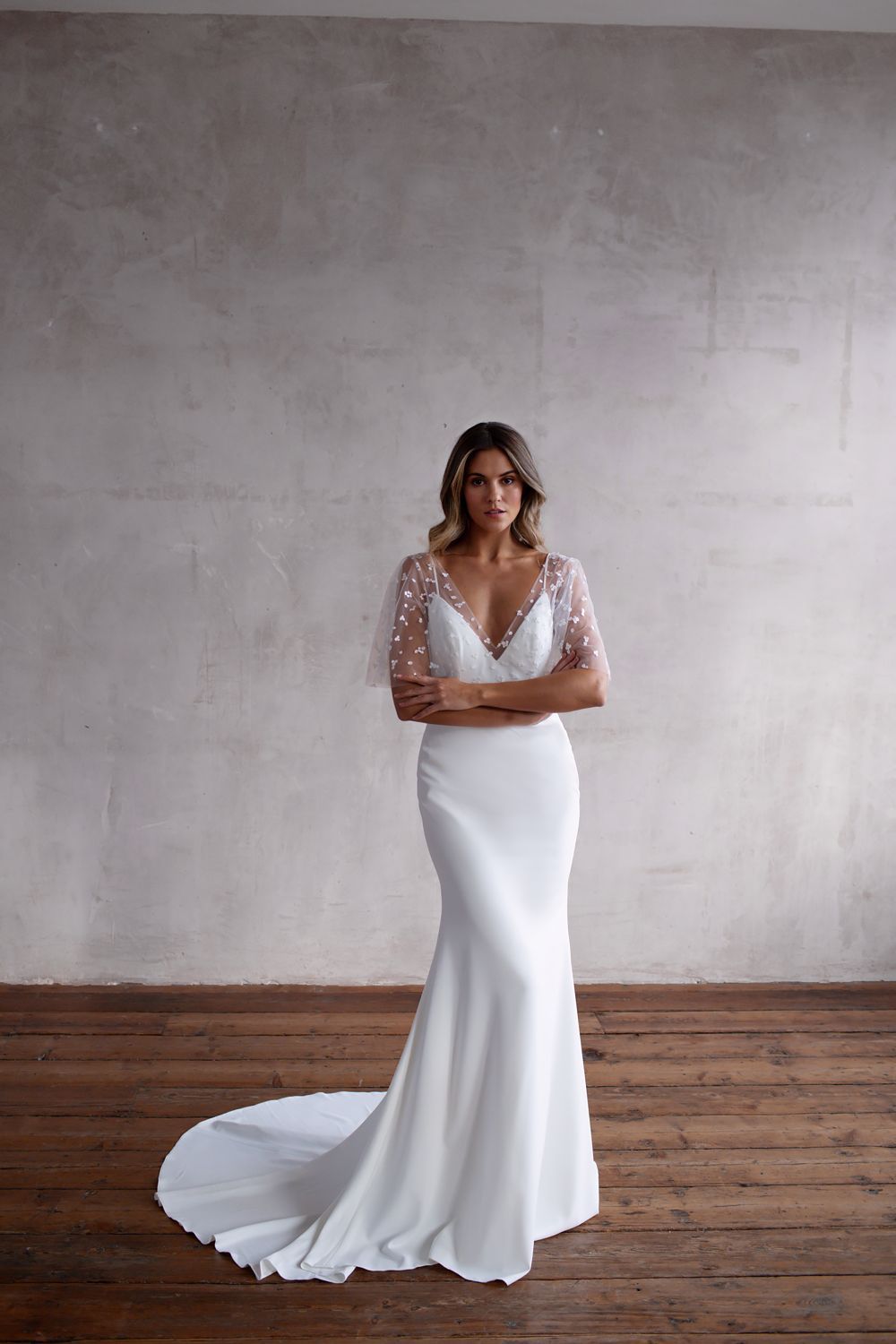 A woman stands against a grey wall wearing a white, floor-length gown with embellished flutter sleeves and a train.