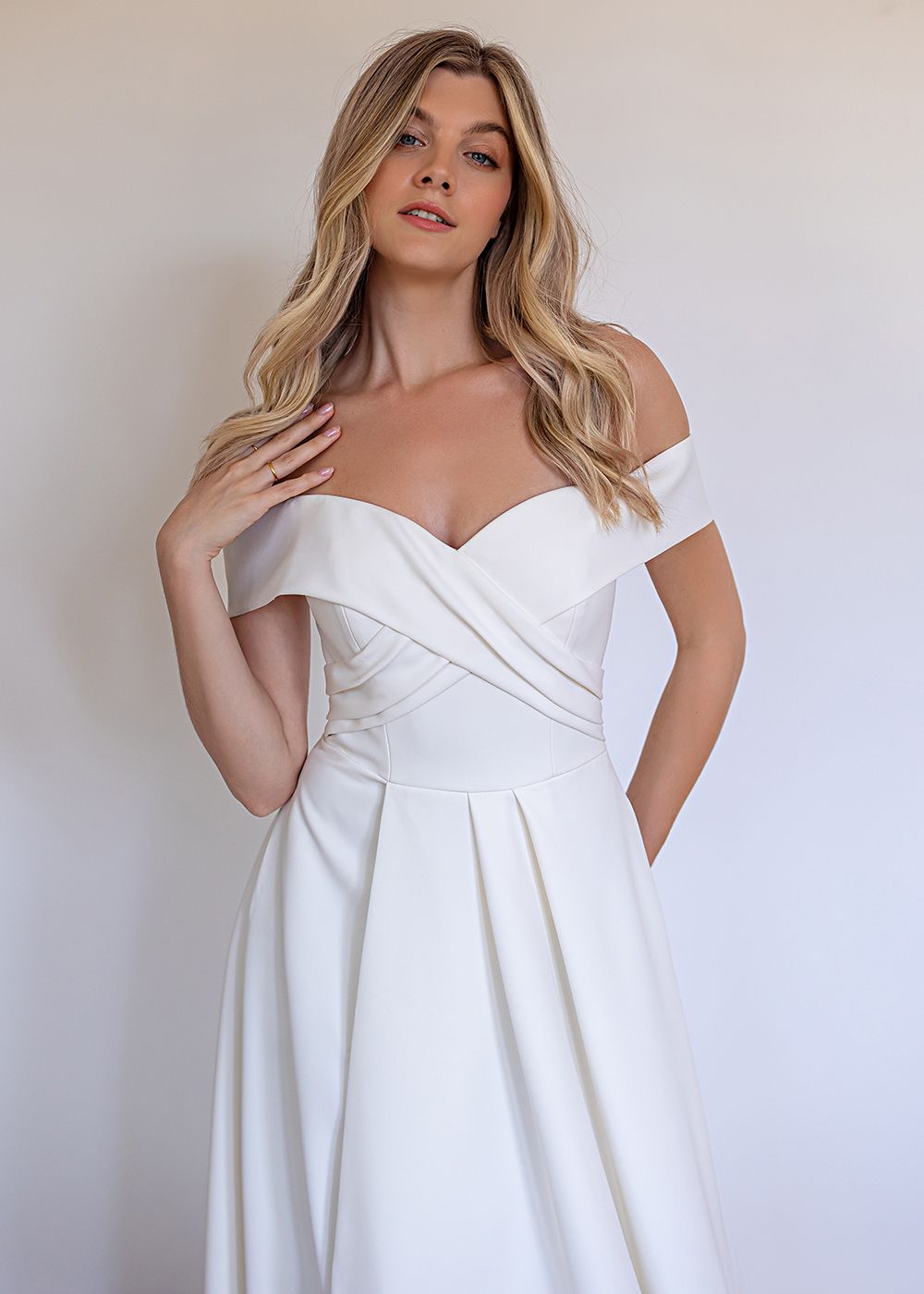 A person wearing a white, off-the-shoulder wedding dress with a crossover bodice and pleated skirt against a plain wall.