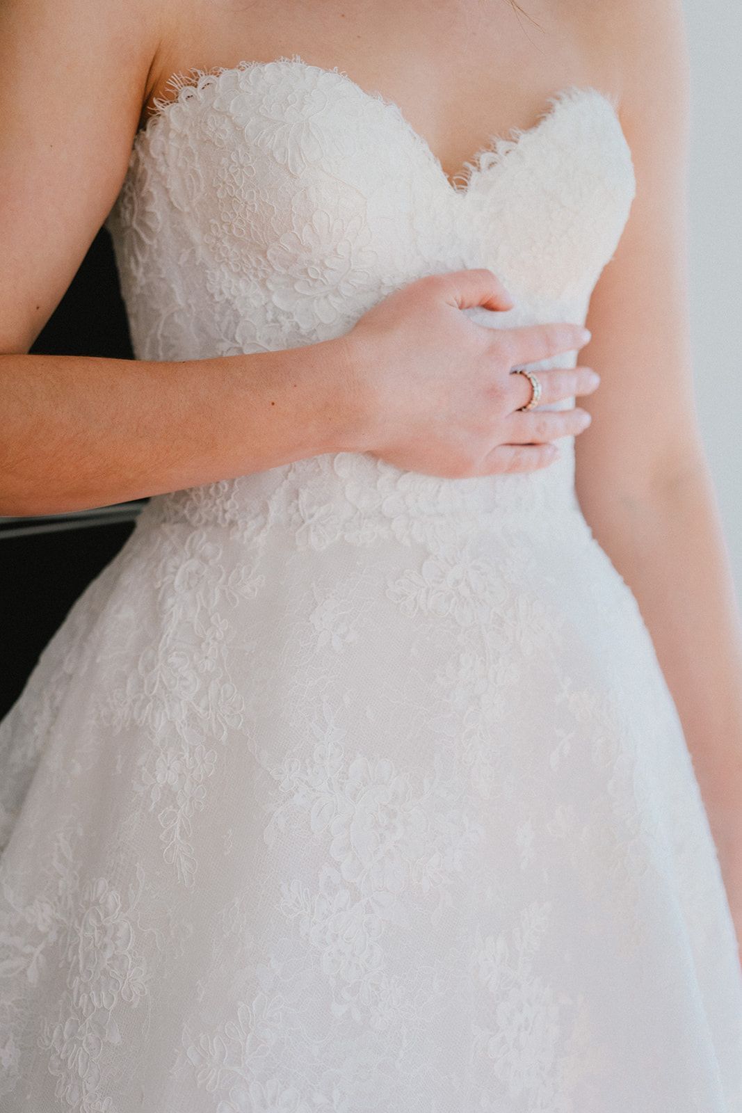 A person wearing a white, strapless, lace wedding dress with their hand resting gently on their waist.
