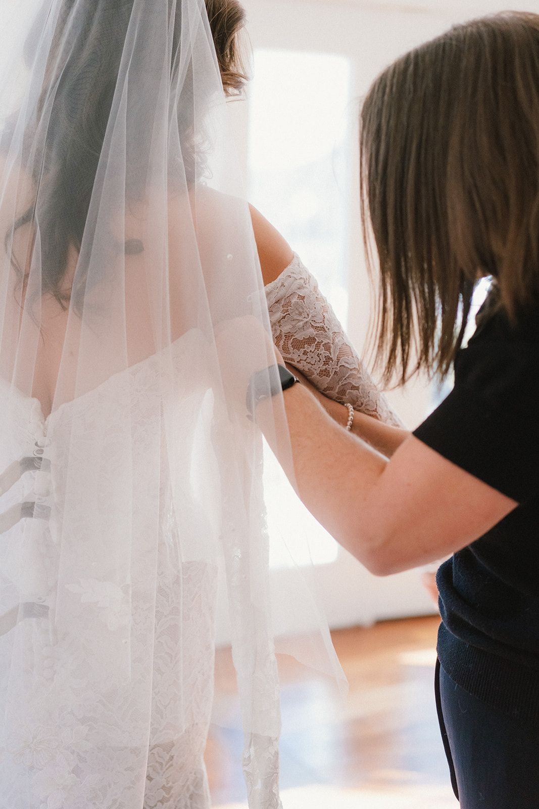 A person in a black shirt helps a bride with her lace-sleeved wedding dress and veil.