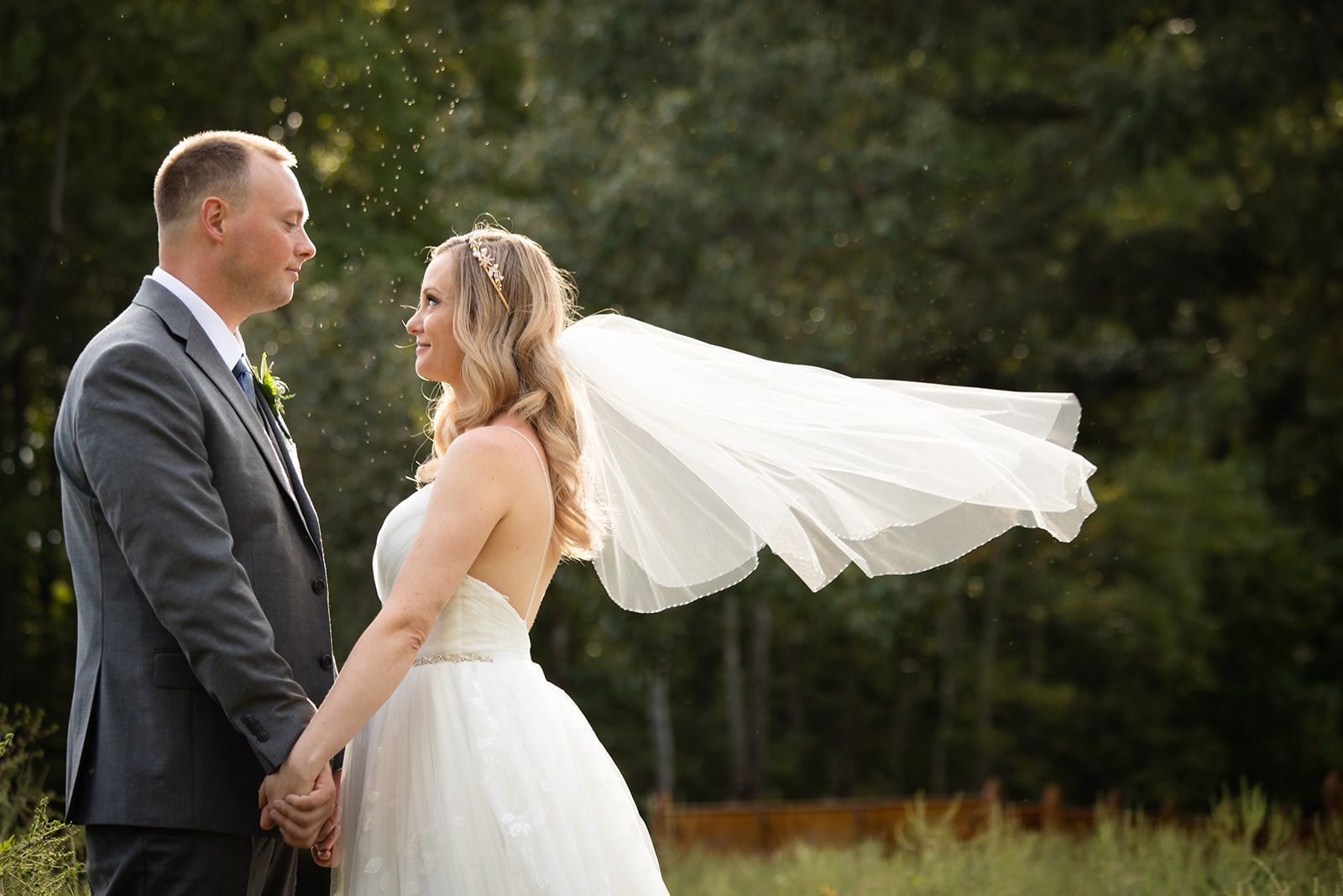 A bride and groom face each other in an outdoor setting with the bride’s veil blowing in the wind.
