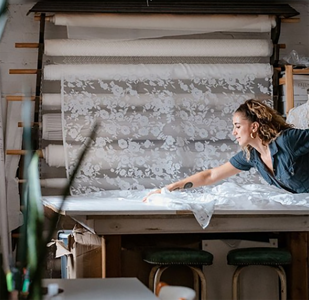 A person working with white floral lace fabric on a wooden table in a studio with multiple rolls of fabric in the back.