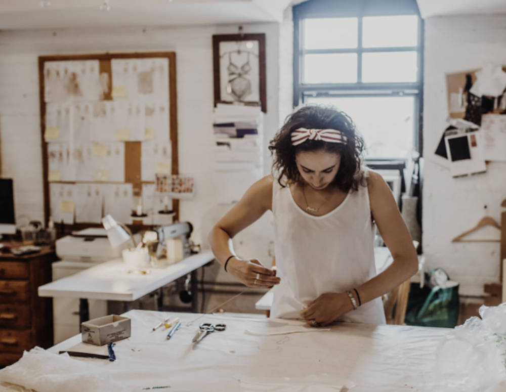 A person wearing a white tank top and headband works on fabric at a desk in a well-lit design studio.