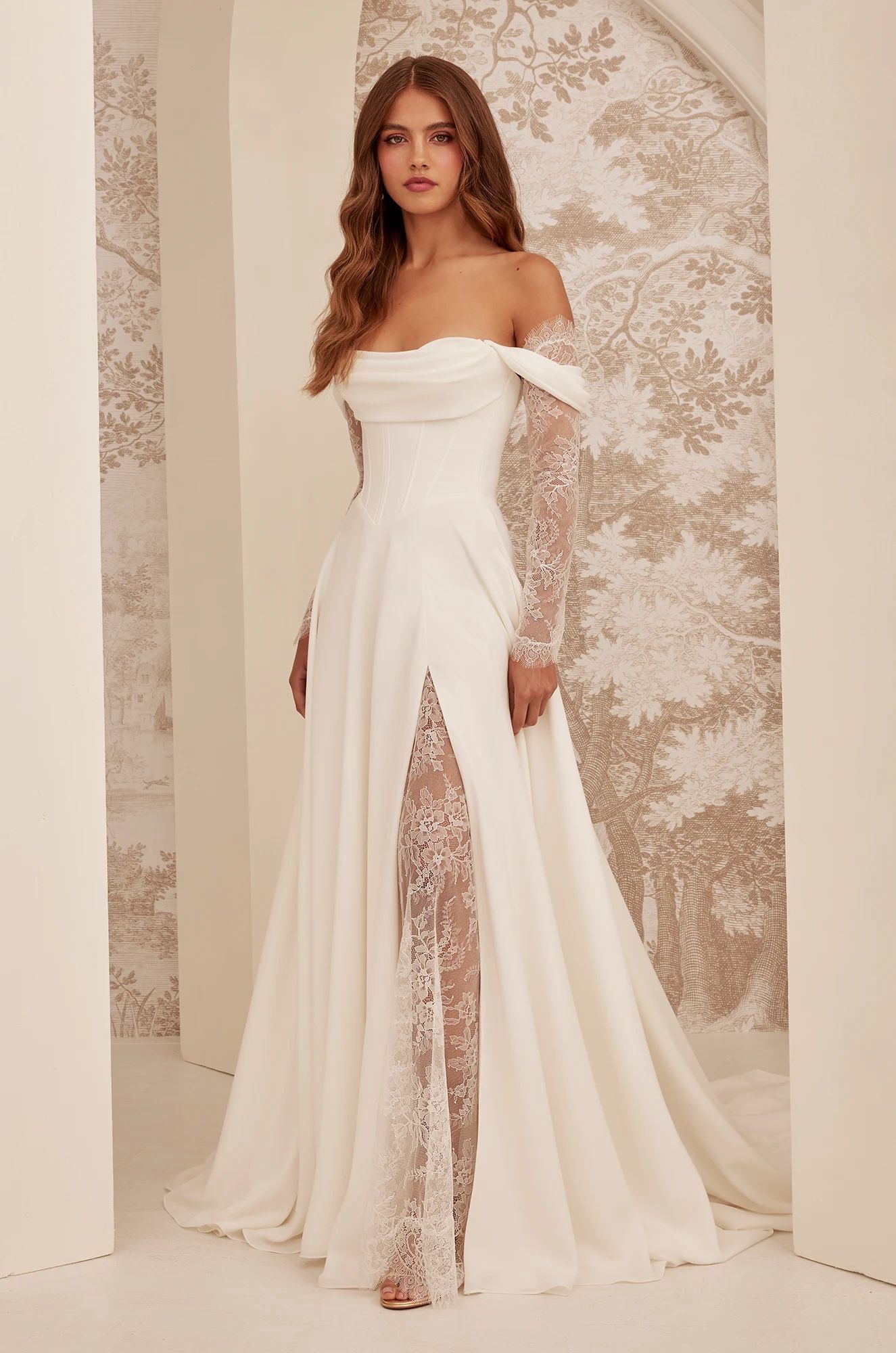 A model wears an off-the-shoulder white wedding gown with long lace sleeves and a high lace-lined leg slit.